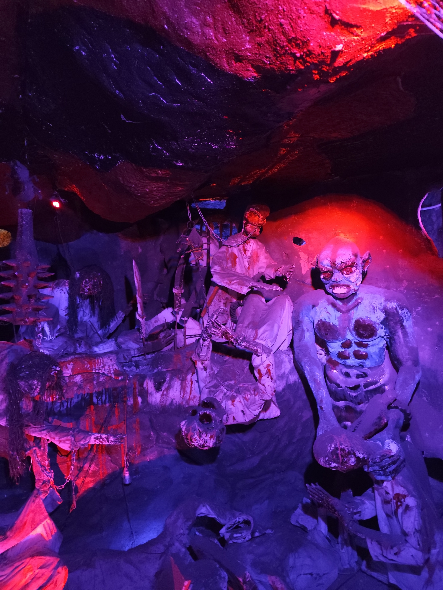 The image depicts a disturbing and gruesome scene, likely a horror display or haunted house attraction. The location is stated as Unknown, Unknown.
Several grotesque figures are present, possibly mannequins or statues. One is a blue-skinned, muscular creature with a sinister expression, standing upright and holding severed limbs. Other figures appear to be victims of violence, with bloodstains and visible wounds. One is hanging by a chain.
The setting appears to be a cave or dungeon-like environment, with rough walls and uneven surfaces. The lighting is dramatic, with a combination of red and purple hues, enhancing the unsettling atmosphere. There are implements of torture or weaponry present, such as spiked objects and blades, adding to the horror theme. The overall scene suggests a place of torment and death.