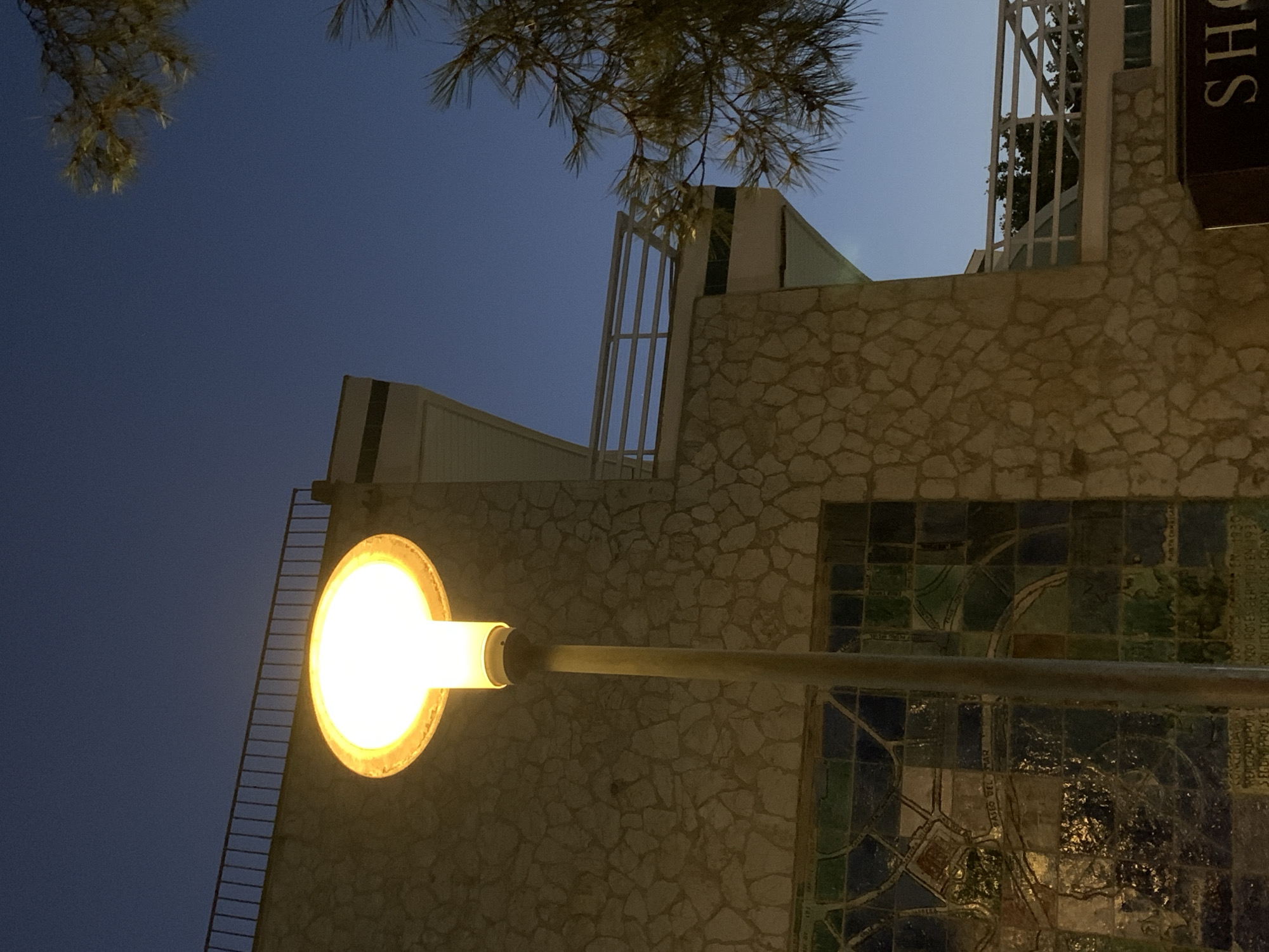 The image shows a street lamp illuminating the exterior of a building with a stone facade and a balcony. The sky is dark blue, suggesting it is dusk or night. A tree with pine needles is visible in the upper right corner. Part of a mosaic is visible on the lower left side of the building, depicting a map-like design and text, potentially indicating a historical or informational mural. A dark sign with the partial word "SH..." is visible in the lower right corner. The overall scene depicts an outdoor urban setting, likely in Palmanova, Spain, given the context provided. There are no people or animals visible in the image.
