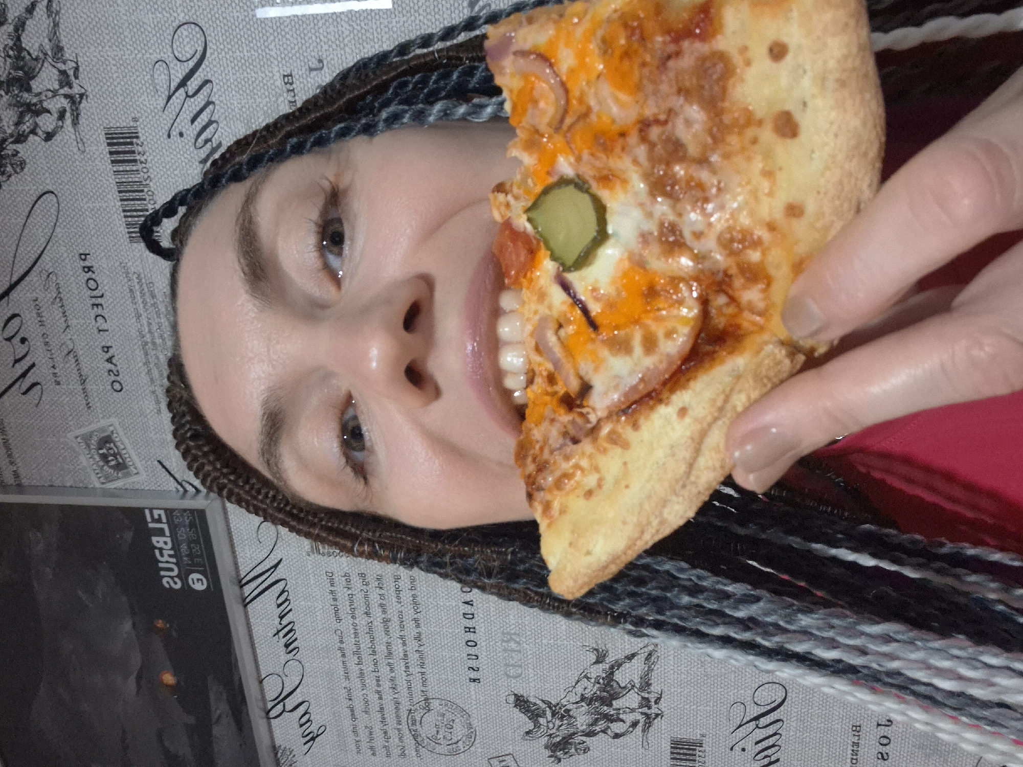 A woman with braided hair is holding a slice of pizza up to her face, obscuring most of it. Her eyes and nose are visible, and she appears to be smiling. The pizza has cheese, red sauce, and what looks like pickles and onions. The background is a textured surface with a repeating pattern of script text and bar codes. The overall impression is a playful and informal moment. Visible text includes various Cyrillic and Latin characters, as well as bar codes and numbers.