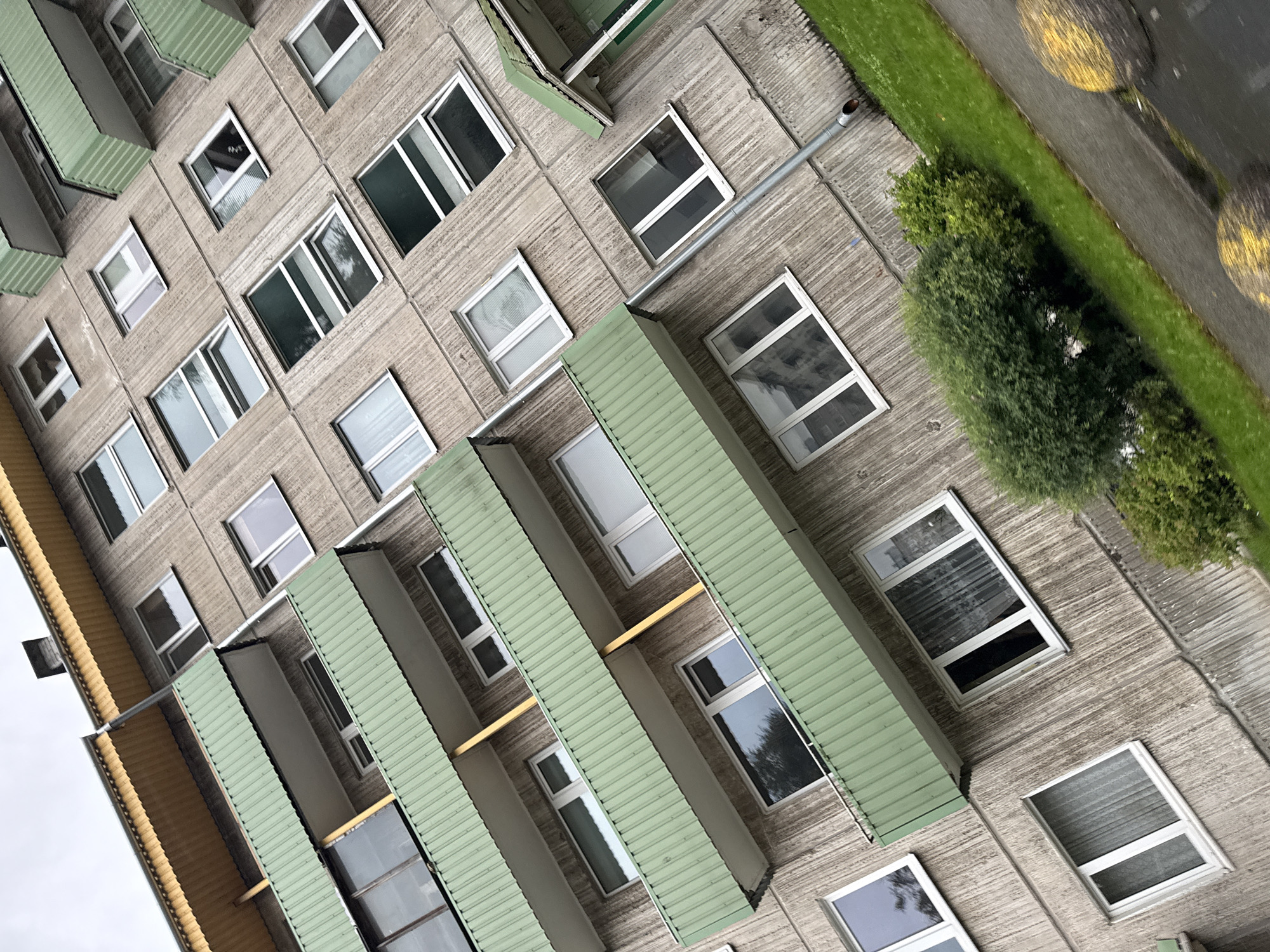 This is an angled, eye-level, outdoor, daylight shot of the facade of a Brutalist apartment building in Tallinn, Estonia. The building is characterized by its concrete construction and the repetition of modular elements. Multiple floors are visible, each with a series of windows and balconies. The balconies are a distinctive pale green color with corrugated metal siding and white railings. The windows are mostly rectangular and appear to be double-paned with white frames. Some windows have blinds or curtains visible. There is a visible drainpipe running vertically down the side of the building. Below the building, a strip of green grass is visible, with two yellow circular markings on a paved surface that appears to be a road or sidewalk. The sky is overcast and a pale gray. There are no people visible in the image.