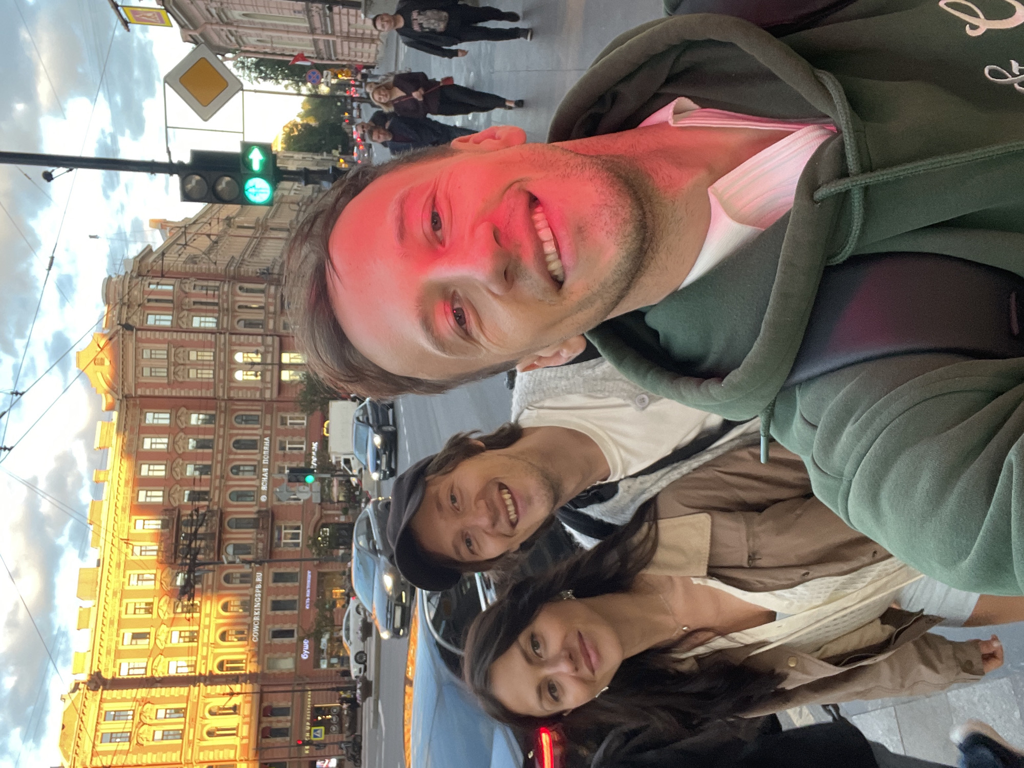 In a street scene in Saint Petersburg, Russia, a group of three individuals are posing for a selfie. The man in the foreground, lit by red light, smiles widely. To his left, a woman with dark hair smiles gently, wearing a light-colored top and a brown jacket. Behind them, a man in a black cap grins, wearing a white t-shirt and a grey cardigan. In the background, pedestrians walk along a sidewalk lined with buildings. A traffic light shows green for pedestrians and a right arrow. Cars are visible on the street. The architecture suggests a historic European city, consistent with Saint Petersburg.