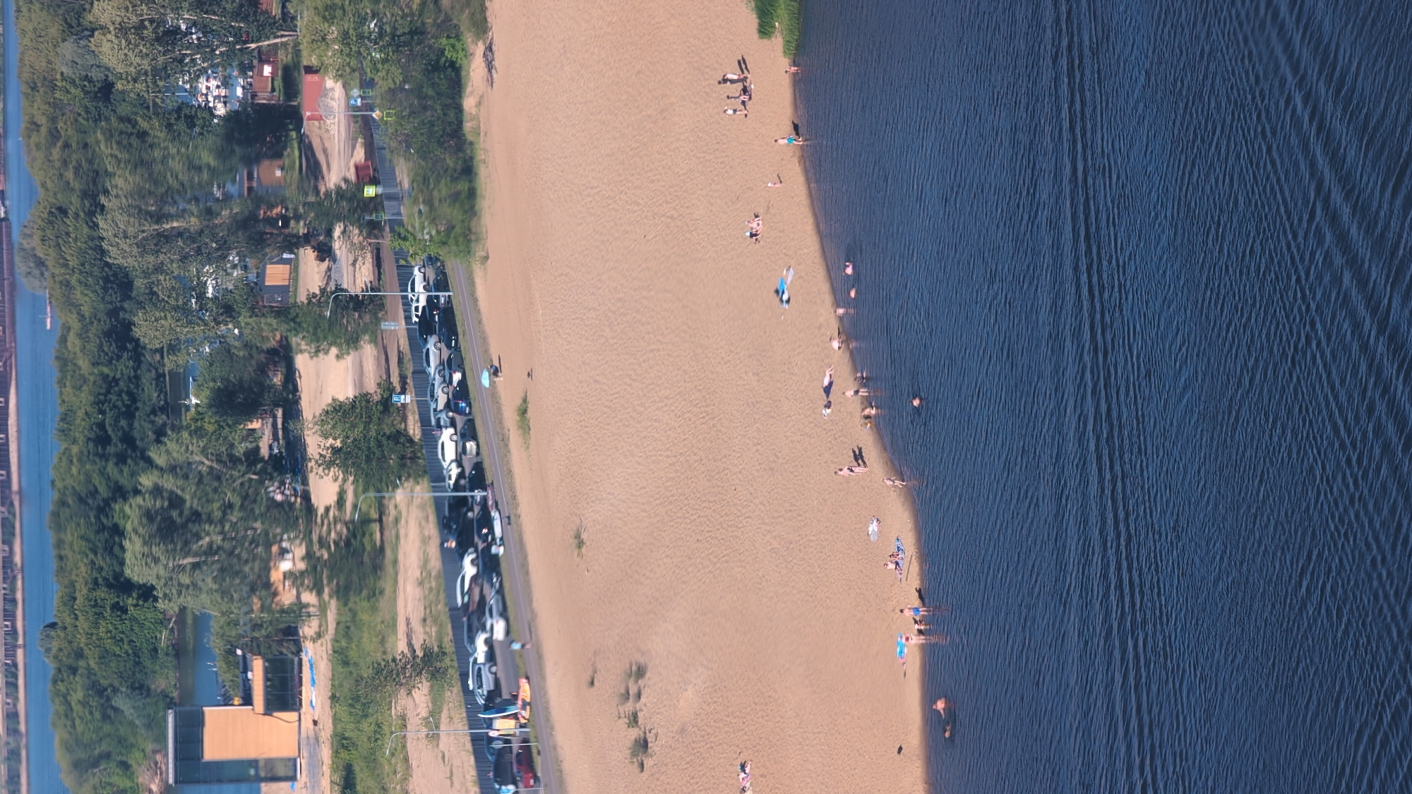 This aerial, wide-angle shot captures a sunlit beach scene on a clear day. The image is oriented vertically. On the left, a dense line of green trees borders a body of dark blue water, hinting at a river or lake. Adjacent to the trees, a paved road is lined with parked cars, mostly white, suggesting a popular recreational area. Further left, glimpses of buildings and more trees are visible.
The central and right portions of the image showcase a sandy beach populated by numerous people. They are scattered across the sand and in the shallow water. The activities observed include sunbathing, swimming, and walking along the shoreline. The sun appears to be relatively high in the sky, casting distinct shadows, indicating it's likely midday or early afternoon. The water's surface is rippled, suggesting a gentle breeze.
The location is identified as Nizhny Novgorod, Russia. While specific emotions are difficult to discern from this aerial perspective, the presence of many people enjoying the water and beach suggests a relaxed and leisurely atmosphere on a pleasant day. No visible text is present in the image.