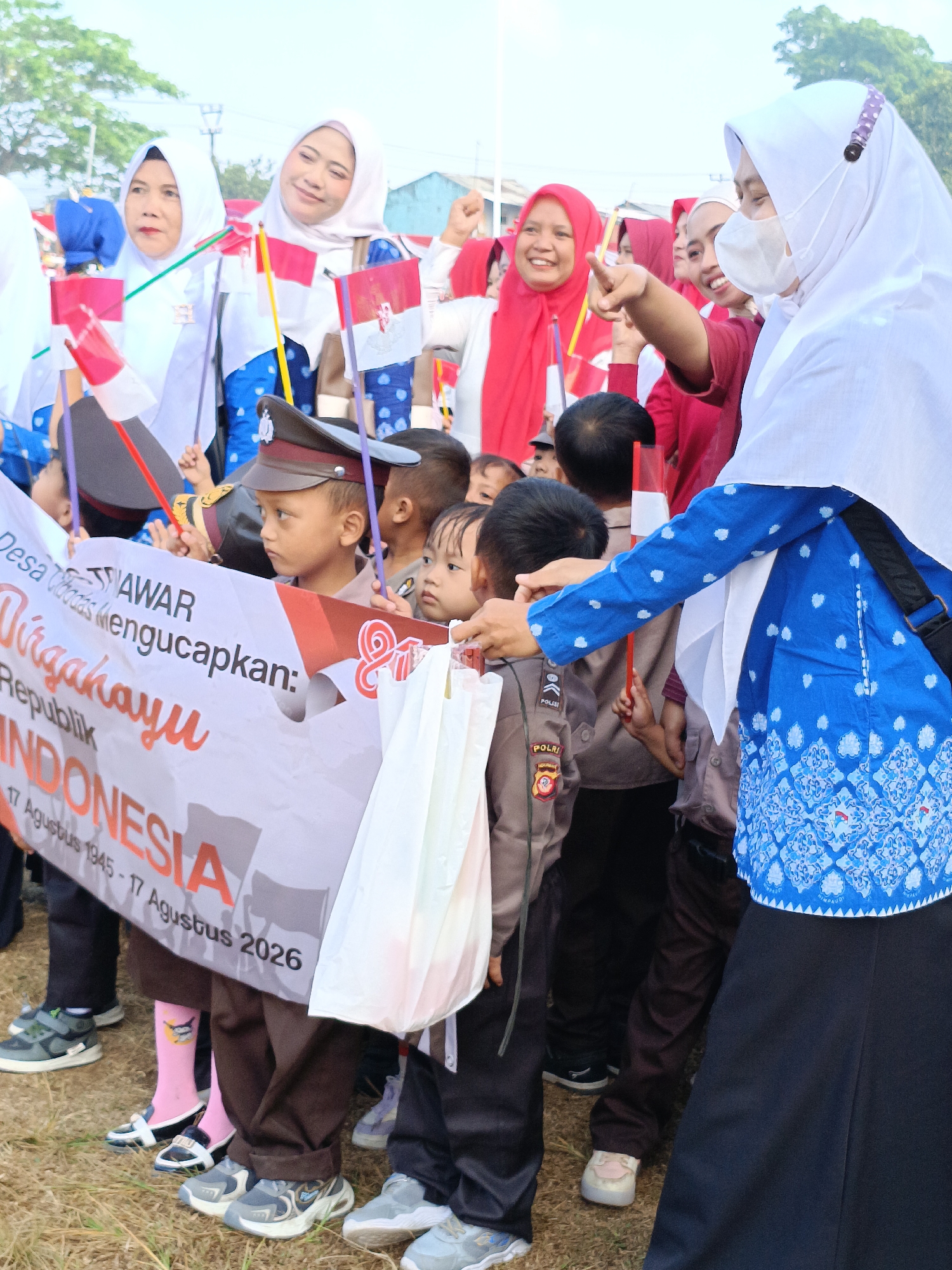 The image depicts a group of people, including children, gathered outdoors in what appears to be a public space, likely during a festive or commemorative event. The presence of Indonesian flags, a banner with text in Indonesian, and the attire of some individuals suggest a national celebration, possibly Independence Day. A banner reads "Dirgahayu Republik INDONESIA" which translates to "Long Live the Republic of Indonesia," further supporting this. Several individuals are wearing traditional Indonesian attire, including headscarves (hijab), and some children are dressed in what appear to be police or scout uniforms. The children are holding small Indonesian flags. One woman, wearing a white hijab and a blue patterned top, is pointing forward. The ground is covered in dry grass, indicating an outdoor setting. The background shows some buildings and trees, suggesting an urban or suburban environment. The event seems to be taking place in Cibodas, Indonesia, as indicated by the banner.