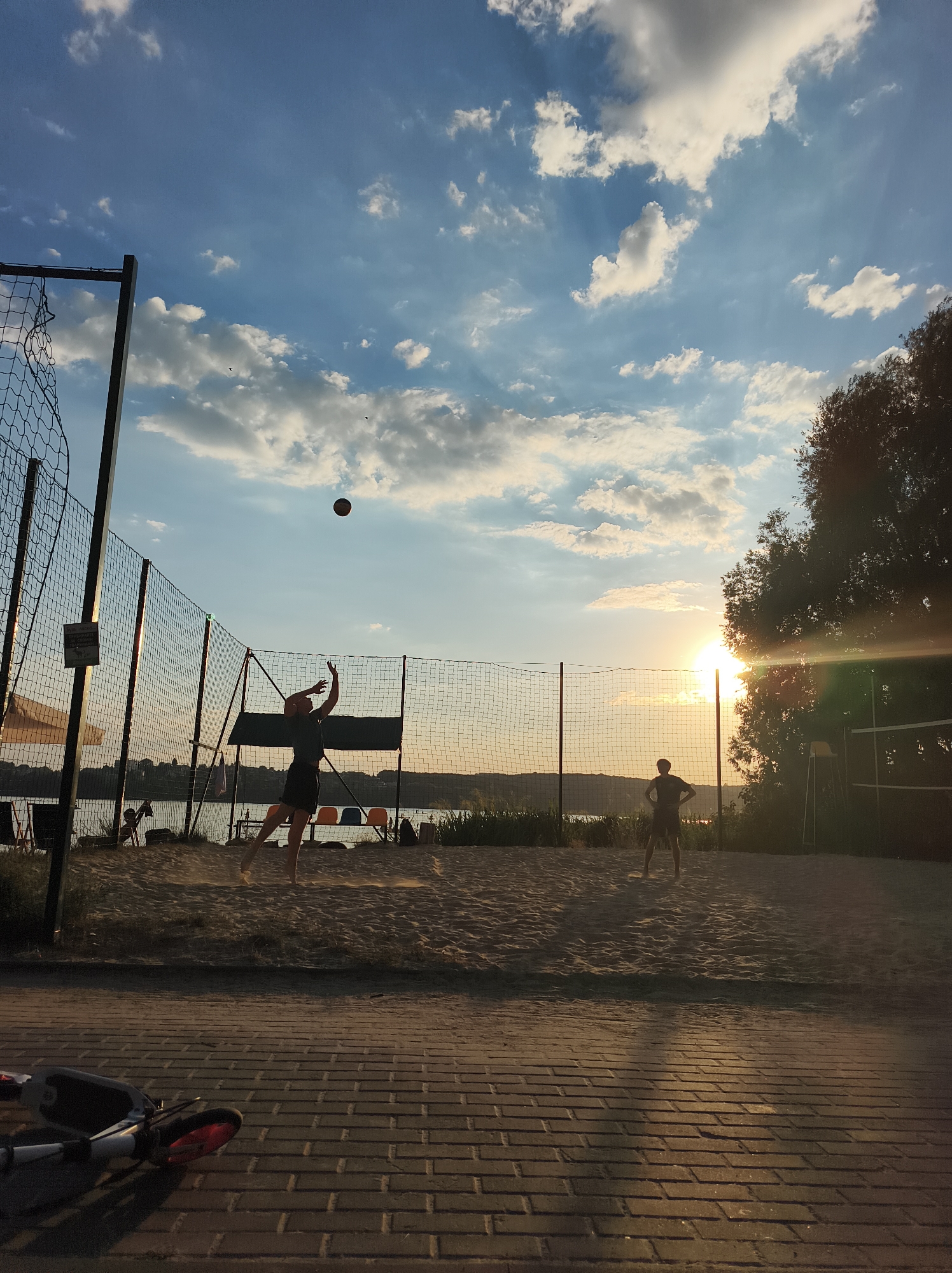 The image depicts a beach volleyball court at sunset. Two individuals are silhouetted against a bright sky, one in the act of serving a volleyball and the other standing on the opposite side of the net. The court is made of sand, and a volleyball net stretches across the middle. In the background, a body of water is visible, suggesting a lakeside or riverside location. Trees line the edge of the water, and distant buildings can be seen. The sky is filled with dramatic clouds, illuminated by the setting sun, which casts a warm glow over the scene. The foreground features a paved area with a scooter lying on its side. The overall atmosphere is serene and sporty, capturing a moment of outdoor recreation.