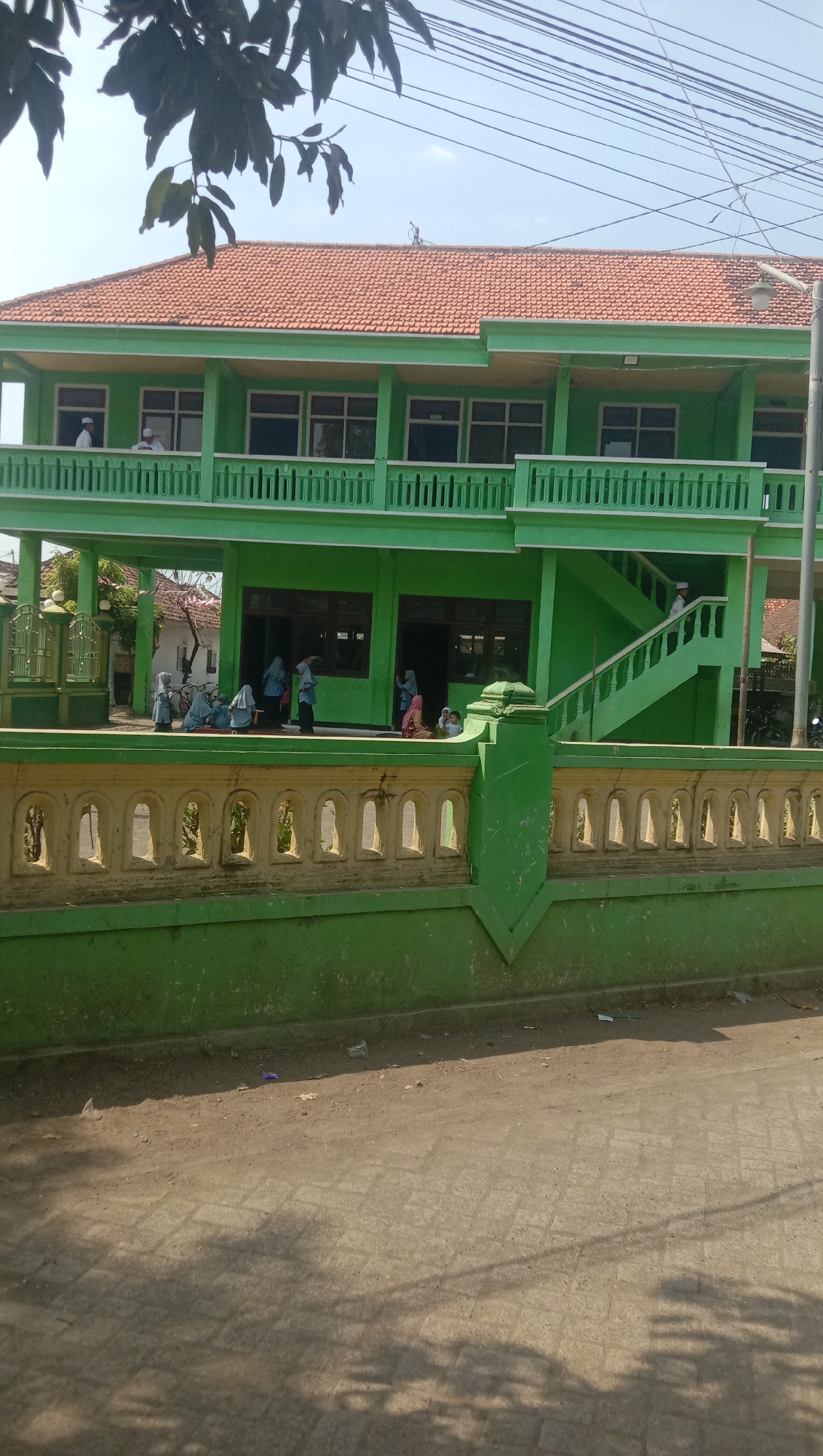 The image captures a bright, sunny day in Ngempit, Indonesia, focused on a two-story green building with a terracotta tile roof. The building has multiple windows and a balcony on the second floor, which is enclosed by a green railing with a decorative pattern. A set of green stairs leads up to the second floor. In front of the building is a low green wall topped with a yellow decorative element featuring arched openings. Several people, appearing to be children and adults, are visible on the ground floor and balcony. Some are wearing traditional Islamic attire, including headscarves. Two individuals in white shirts and caps are on the second-floor balcony, near the stairs. On the ground floor, a group of individuals, some wearing hijabs, are gathered near the entrance and on a small porch area. One person is seen walking up the stairs. The area in front of the wall is a dirt path with some paving stones visible, and shadows from trees are cast across it. The overall scene suggests a religious or educational institution, possibly a mosque or school, in a rural or semi-rural setting.
