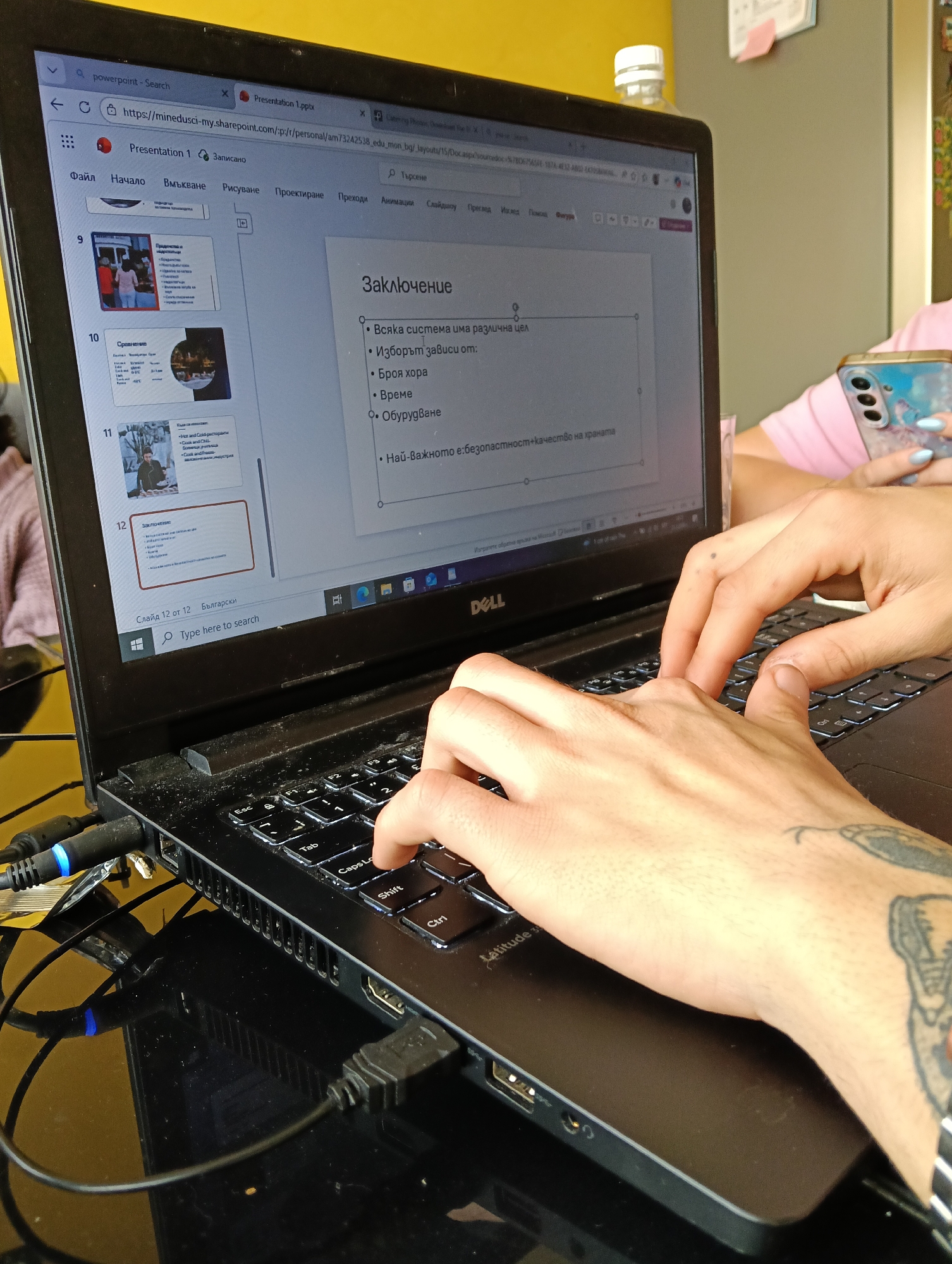The image shows a person typing on a Dell Latitude 3 laptop. The laptop screen displays a PowerPoint presentation in Bulgarian, with the 12th slide visible, titled "Заключение" (Conclusion). The slide lists bullet points about the system having different purposes and the selection depending on factors like the number of people, time, and equipment, with the main point being safety and quality of food. The laptop is connected to external devices via USB and HDMI cables. A woman with a pink shirt and her phone is in the background. Based on the PowerPoint being in Bulgarian, the location is in Bulgaria.