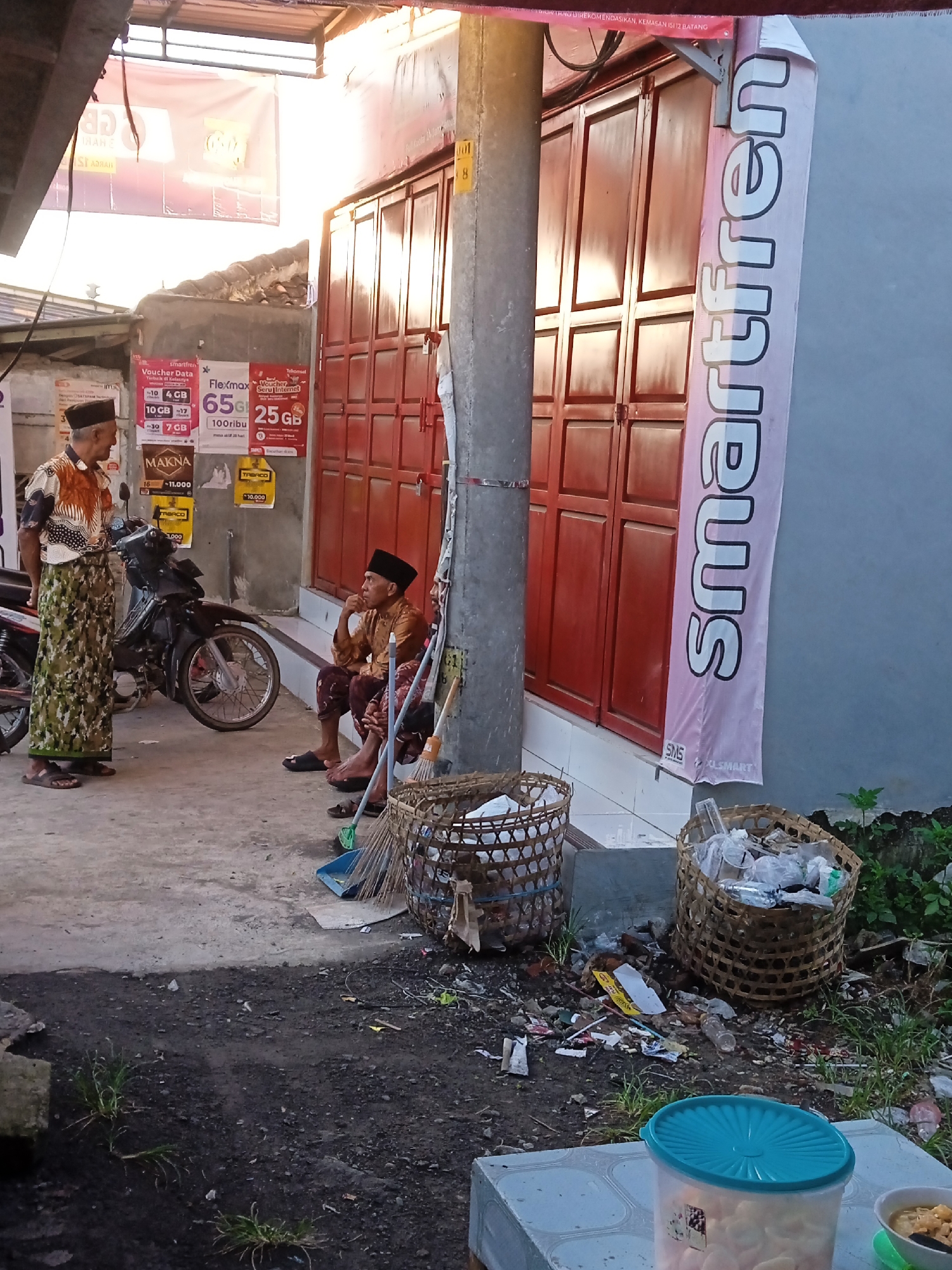 The image depicts a street scene in Sidorejo, Indonesia. Two older men wearing traditional Muslim attire, including skullcaps, are present. One man stands in a printed shirt and sarong, while the other sits, dressed in a brown long-sleeved shirt and sarong. A motorcycle is parked nearby. Behind them, a red shuttered door is visible, adorned with advertisements for "Smartfren" and data vouchers. The ground is concrete, with a small pile of litter and debris in the foreground, including two woven baskets filled with waste. A blue-lidded plastic container sits on a tile surface in the foreground. A utility pole is positioned in the center.
