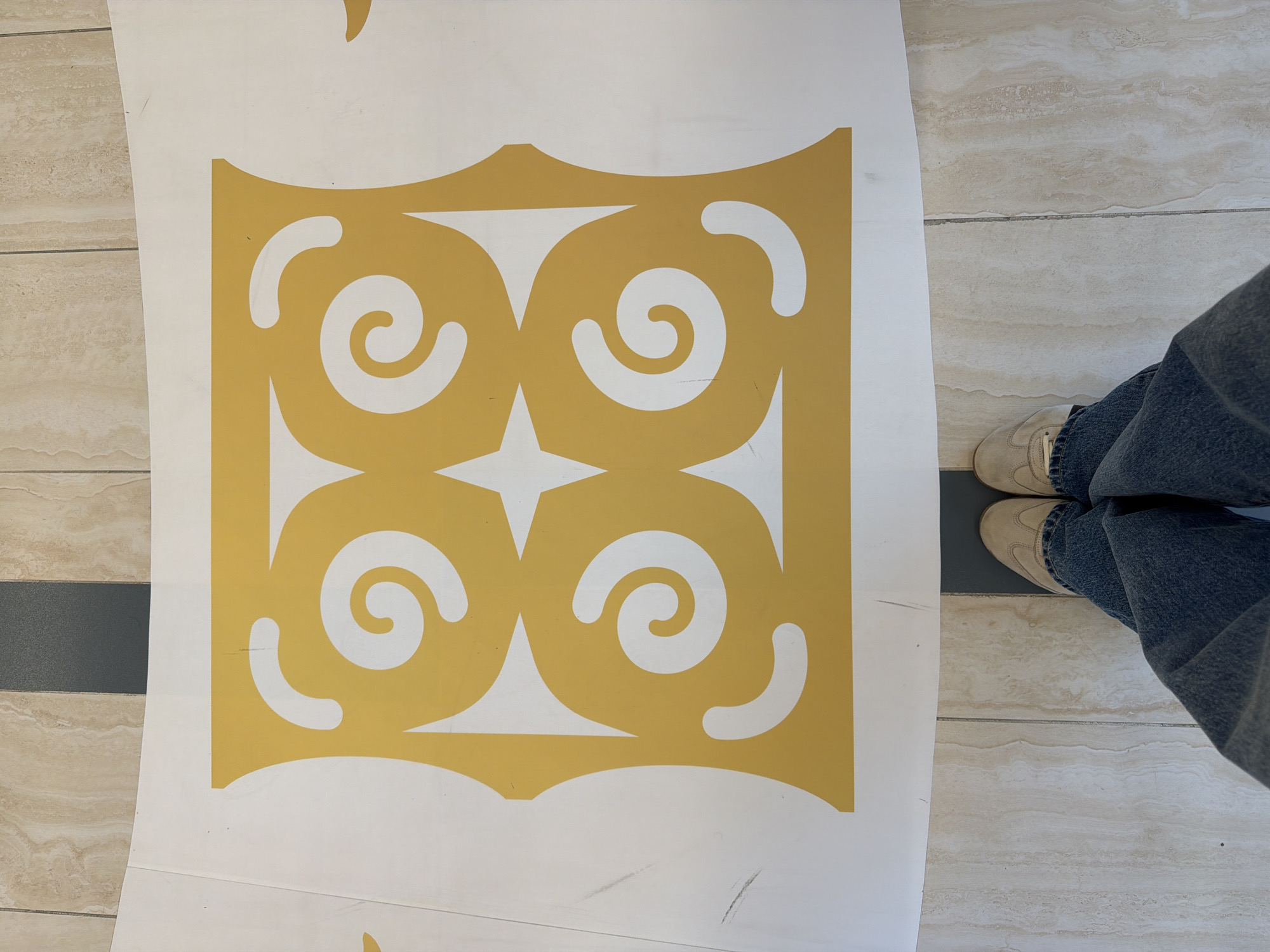 The image shows a close-up, overhead view of a person's feet wearing blue jeans and tan shoes, standing on a tiled floor. The feet are positioned in front of a large white sheet with a yellow geometric pattern. The pattern features four large, white, stylized spiral shapes arranged in a square, with a white star shape in the center. The floor tiles are a light beige with subtle veining. A dark gray strip runs across the floor behind the white sheet. The scene appears to be indoors, possibly in a public space or a room with a large graphic element. There is no indication of the location being Yuzhno-Sakhalinsk, Russia.
