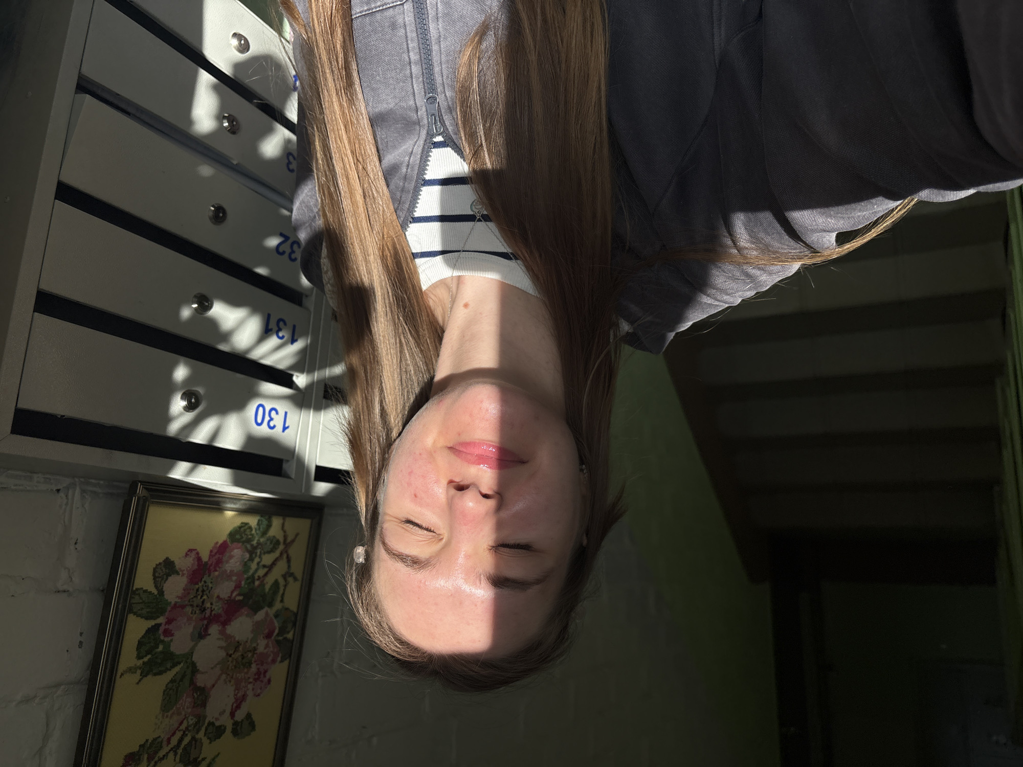 A photograph shows a fair-skinned person with long, light brown hair, wearing a striped shirt and a gray jacket, with their head upside down in a room. The person's eyes are closed. Part of their face is illuminated by direct sunlight. To the left of the person are mailboxes labeled with numbers including "130", "131", "32", and "3." A framed floral embroidery hangs below the mailboxes. To the right, there is an entryway with dark green walls and stairs leading upwards into deeper shadow. The image was taken indoors. The location is Perm, Russia.