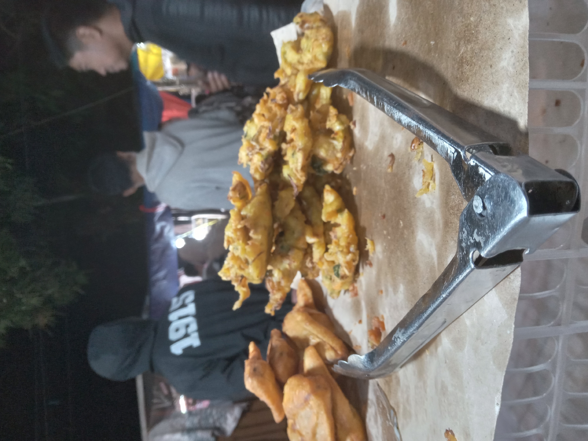 In an outdoor, nighttime setting, a close-up shot focuses on a spread of fried food items, possibly fritters or pakoras, stacked on a light brown surface. A pair of silver metal tongs rests in the foreground, open and positioned over the food. Behind the food, three individuals are visible, mostly from the back. The person on the far left wears a black hooded sweatshirt with "1915" printed in white on the back. To their right, another person wears a light gray hooded sweatshirt. The person on the far right is seen from the side and wears a dark jacket. The background is dark and appears to have some stalls or tents with lights, suggesting a market or food stall environment. The overall lighting is dim, with some harsh illumination from above, contributing to the nighttime ambiance.