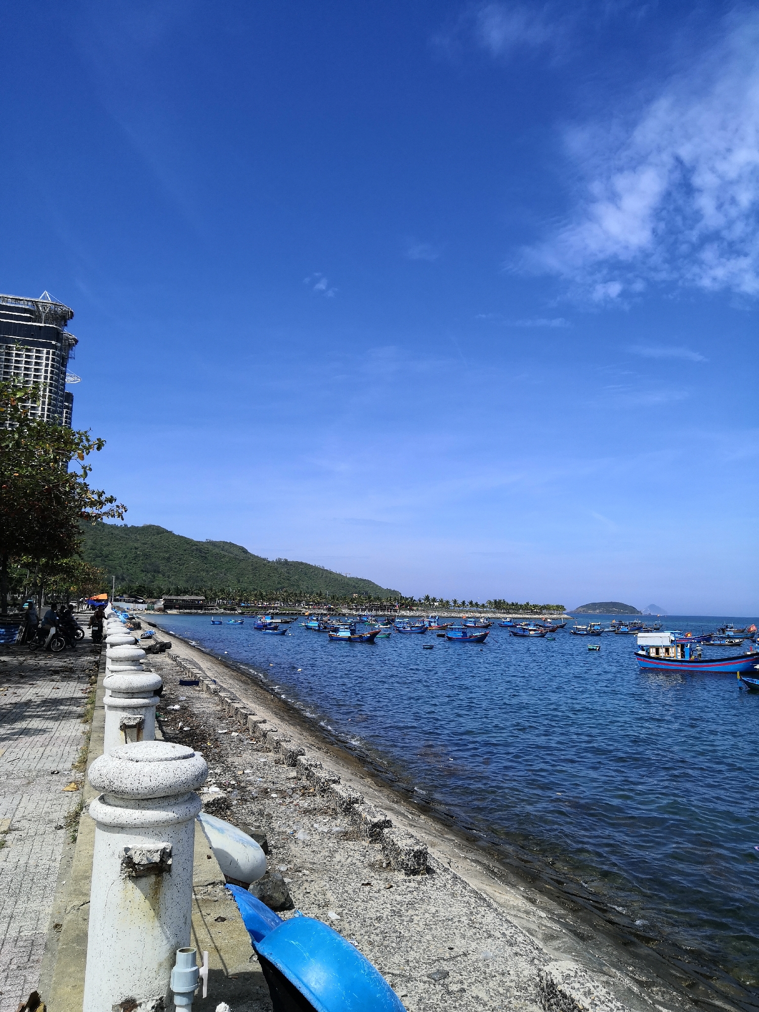 The image captures a coastal scene in Bac Nha Trang Ward, Vietnam, on a clear day with a bright blue sky and sparse white clouds. In the foreground, a textured, gray concrete promenade lines the waterfront, demarcated by a series of white, cylindrical stone bollards. To the left of the promenade, a few individuals are seen near motorcycles, suggesting a public walkway or parking area. Adjacent to the promenade, a tree with green foliage stands on the left.
The shoreline itself is a mix of rough, gray concrete and a stony, debris-strewn edge. Beyond the shoreline, the deep blue sea is dotted with numerous small fishing boats of various colors, predominantly blue, white, and some with red accents. A larger, more detailed blue and white boat with a cabin is prominently positioned towards the right side of the image. In the background, a lush green, treed mountain range forms the backdrop to the coastal town, which is characterized by low-rise buildings and palm trees along the water's edge. A tall, unfinished building with a visible scaffolding structure is situated on the far left, partially obscuring the base of the mountain. The overall impression is one of a bustling yet serene coastal community.