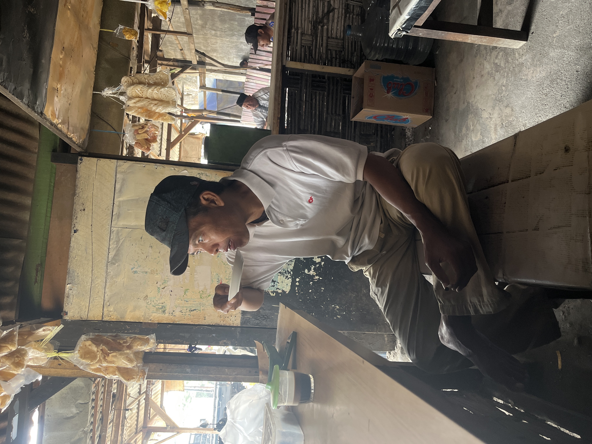 The image depicts a man wearing a white polo shirt and khaki pants, seated on a wooden bench, drinking from a small white cup. He has a dark baseball cap on and is looking to his left. The setting appears to be an open-air market or roadside stall in Dusun Metatu, Indonesia. Hanging bags of snacks are visible overhead. A wooden table with a glass of dark liquid and other items is to the man's left. In the background, other individuals are visible, and there are large water jugs and a cardboard box on the floor. The overall environment suggests a casual, possibly commercial, outdoor setting.