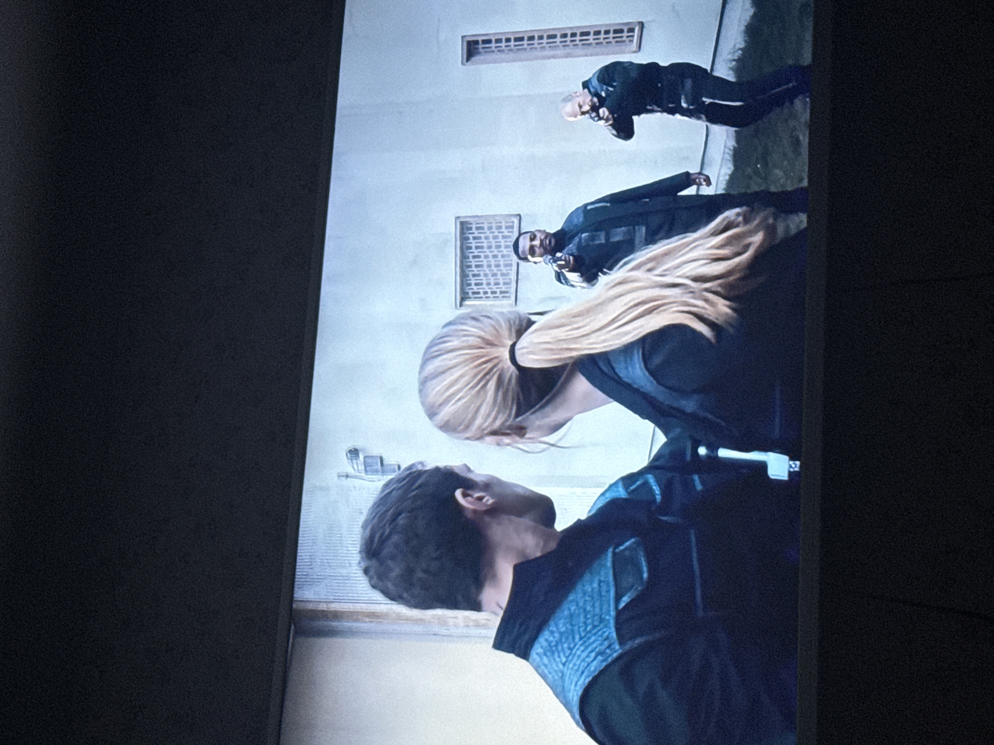 The image depicts a scene with three people. Two are positioned in the foreground, appearing to be close to each other. One has short dark hair, while the other has long blonde hair pulled back in a ponytail. Both are wearing dark clothing with blue accents. In the background, a third person, who is bald, is positioned further away and also wearing dark clothing. The environment consists of a light-colored building exterior with what appears to be ventilation windows. The location is consistent with an outdoor area near a building. The image does not contain sufficient information to determine a specific location within the city of Mytishchi, Russia.