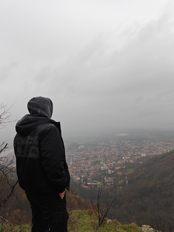 A solitary individual, observed from behind, stands on a rocky, grassy prominence overlooking the city of Shumen, Bulgaria. The person is wearing a dark, possibly black, hooded jacket with the hood raised, obscuring the head, and dark trousers; the right hand is partially inserted into a pocket. Sparse, bare tree branches are visible in the immediate foreground. Below, the city of Shumen extends across the valley, characterized by a dense arrangement of buildings with various roof colors, predominantly dark. The urban area transitions into fields and distant, hazy landscape features, including a light-colored expanse in the far background. The entire scene is under a uniformly overcast, grey sky, indicating low visibility and diffuse lighting conditions.