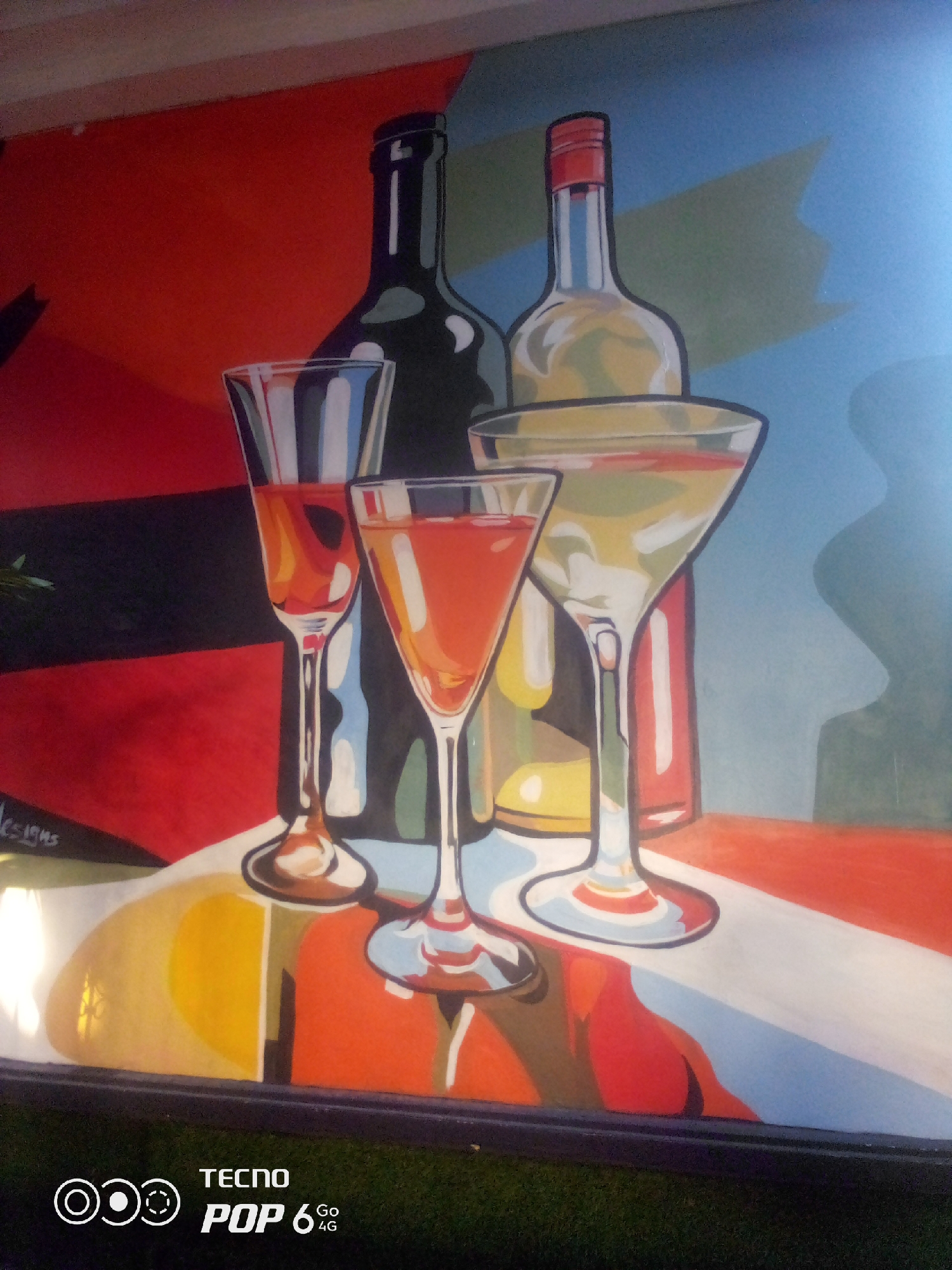 The image shows a brightly colored mural depicting two bottles and four glasses of drinks. The mural features a dark bottle and a lighter, clear bottle with a red cap. Three of the glasses are filled with red or orange liquid, and one martini glass is filled with a pale yellow liquid. The background is a vibrant mix of red, blue, and yellow abstract shapes. A signature reading "Designs" is visible in the lower left corner of the mural. The bottom edge of the image shows a dark frame or border, and below that, grass. The image includes a watermark with "TECNO POP 6 4G" text and a logo. This mural is located in Howayi, Nigeria.