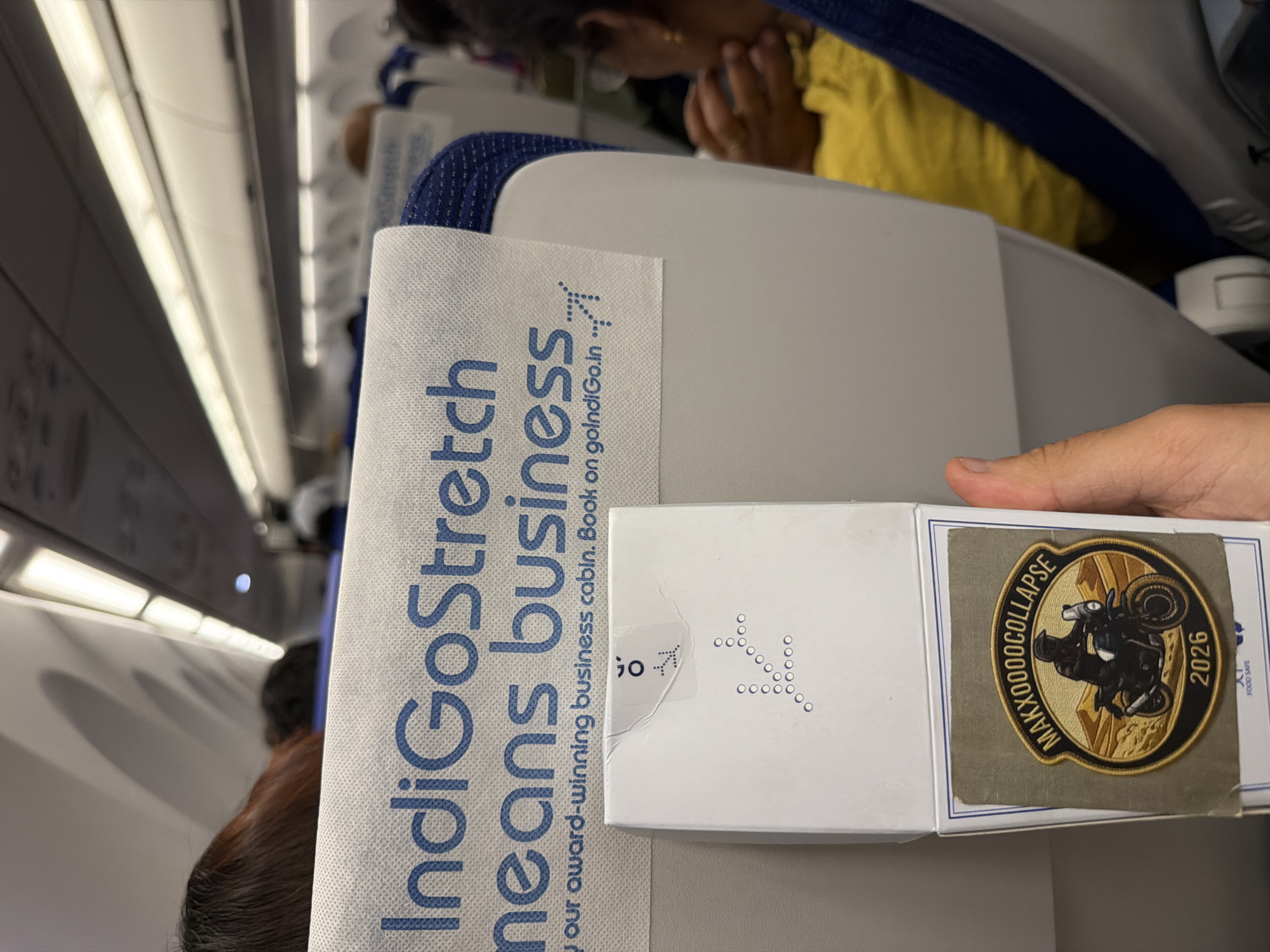 A close-up, eye-level shot shows a hand holding a white box inside an airplane cabin. The box has a circular sticker with the text "MAXXOOOOCOLLAPSE" and the year "2026" printed below an image of a person riding a motorcycle in a desert landscape. The airplane seats have headrests with the Indigo Airlines logo printed in blue. The lighting suggests it is daytime.