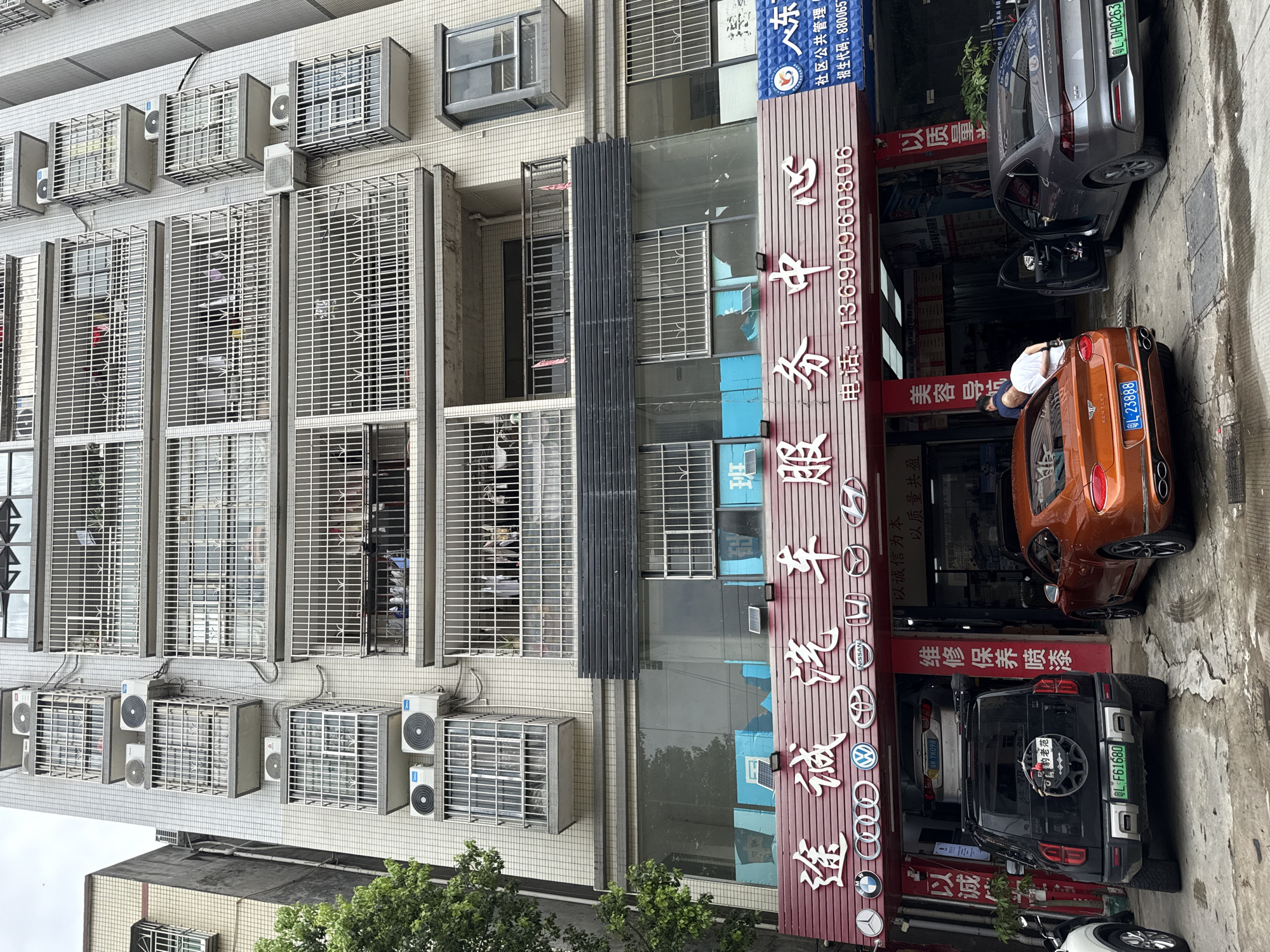 The image shows a street view in Huizhou, China, focused on a multi-story building with a car service center at the ground level. The building's facade is a mix of beige tiled sections and concrete, with numerous windows protected by white metal grates. Multiple air conditioning units are mounted on the exterior walls. The ground floor features a wide storefront with a red awning displaying Chinese characters that translate to "Tu Cheng Car Service Center" and a phone number. Below the main sign, various car brand logos, including BMW, Audi, Volkswagen, Nissan, Honda, and others, are visible.
In front of the service center, several cars are parked. The most prominent is an orange Bentley Continental GT, with its rear facing the camera. To its left is a black SUV, possibly a modified Toyota RAV4 or similar, with a spare tire mounted on the back. To the right of the Bentley, a grey BYD sedan is parked with its driver's side door open. Two individuals are near the Bentley, one appearing to be a mechanic in white clothing and the other in a blue shirt. The ground is a rough concrete surface, with some water and tire marks. The overall environment suggests a commercial area within a Chinese city.