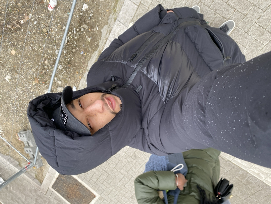 A male subject is depicted in a high-angle, selfie-style shot in Bischheim, France. He is positioned prominently in the foreground, facing the camera with a neutral expression, his gaze directed upward. He has short facial hair, specifically a goatee and a mustache. The subject wears a black puffer jacket with a large hood pulled up, obscuring his ears and part of his head. A dark baseball cap is visible underneath the hood. Numerous small water droplets are visible on the left sleeve and shoulder of his jacket, indicating precipitation.
To the left and slightly behind the primary subject, a second individual is partially visible, oriented perpendicular to the camera's view. This individual wears a green puffer jacket and what appears to be a blue sweater or shirt. A small black handbag is clutched in their left hand, which is also partially visible.
The immediate ground surface beneath the subjects consists of light-colored paving stones. Beyond the paved area, a construction or unkempt dirt/gravel patch is visible. This area contains scattered debris, including small rocks and what appears to be a crushed white disposable cup. A metallic barrier or fence structure, comprising horizontal bars, separates the paved area from the dirt patch. Two light-colored concrete blocks are discernible near the fence in the upper left background. The overall lighting suggests an overcast sky.