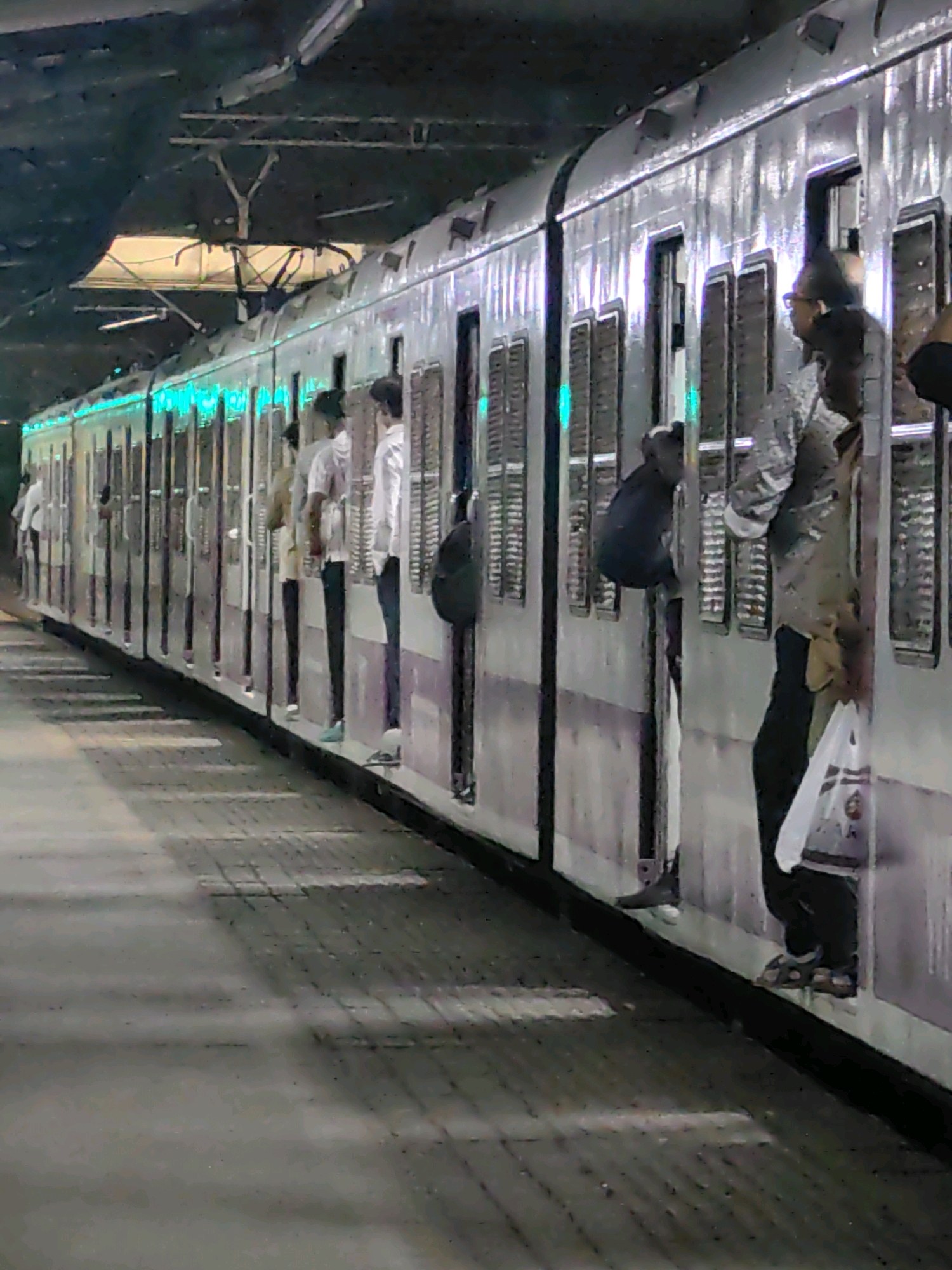 A crowded train car is visible from the outside, alongside a platform in Mira Bhayandar, India. The train is silver with green lights lining the top of the passenger cars. People are leaning out of the open windows of the train, some with their upper bodies visible and others with only their legs. A few individuals are standing on the platform next to the train. The platform is paved with grey tiles and is dimly lit. The overall scene depicts a busy train station with commuters preparing to board or disembark.