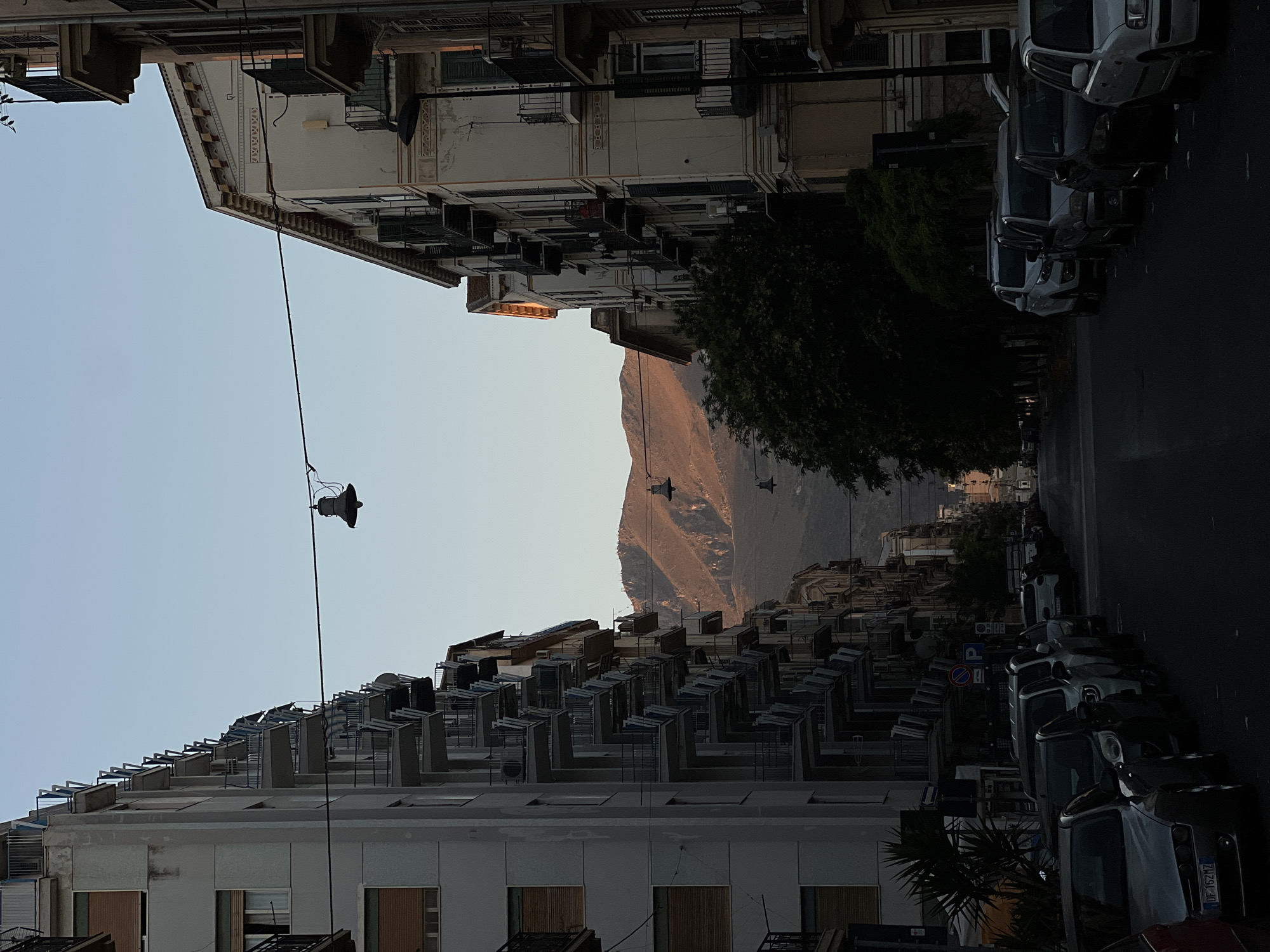 This is a street scene in Palermo, Italy, likely during the late afternoon or early evening. Tall apartment buildings with balconies line both sides of a street. The buildings on the left are gray and have numerous balconies, while the buildings on the right are lighter colored with decorative elements. In the distance, a mountain range is visible, bathed in the warm light of the setting sun. A single lamppost hangs from a wire stretched across the street. The street is lined with parked cars, and the overall atmosphere is one of quiet urban life.