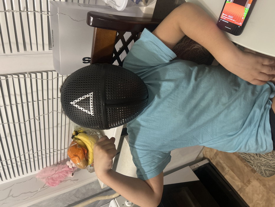An individual, likely a child or young adult, is depicted seated in an indoor setting, wearing a black, perforated mask that covers their entire face. The mask features a prominent white triangular symbol composed of small white dots, a clear reference to the "Squid Game" series. The person is wearing a light blue short-sleeved t-shirt and dark-colored shorts or trousers.
They are seated at a light-colored table, leaning against a patterned cushion on a dark brown wooden chair with a lattice-style backrest. Their right arm rests casually on the table, next to a black smartphone that is lying flat. The phone's screen displays a colorful, pixelated video game, with noticeable red-orange user interface elements, resembling games like Minecraft. The person's left arm is bent, and their hand is near a clear plastic bag containing several yellow bananas and at least one orange-colored fruit, possibly a persimmon or small pumpkin. A fluffy pink decorative object or toy is also visible next to the fruit bag.
In the background, to the person's left, there is a tall, light-colored slatted structure, possibly a radiator cover or a decorative wall panel, across which delicate string lights are draped horizontally. Behind the wooden chair, a white box with a black letter "S" (possibly part of a brand logo) is visible on a surface, along with a white plastic container. In the lower part of the frame, underneath the table and chair, a dark-colored kitchen appliance with a reflective glass door, likely an oven or dishwasher, can be seen, along with a light-colored floor or rug. The scene suggests a relaxed, indoor environment, possibly a kitchen or dining area, with the individual engaged in leisure activities. The time of day is not explicitly indicated, and the mask obscures any facial expressions.