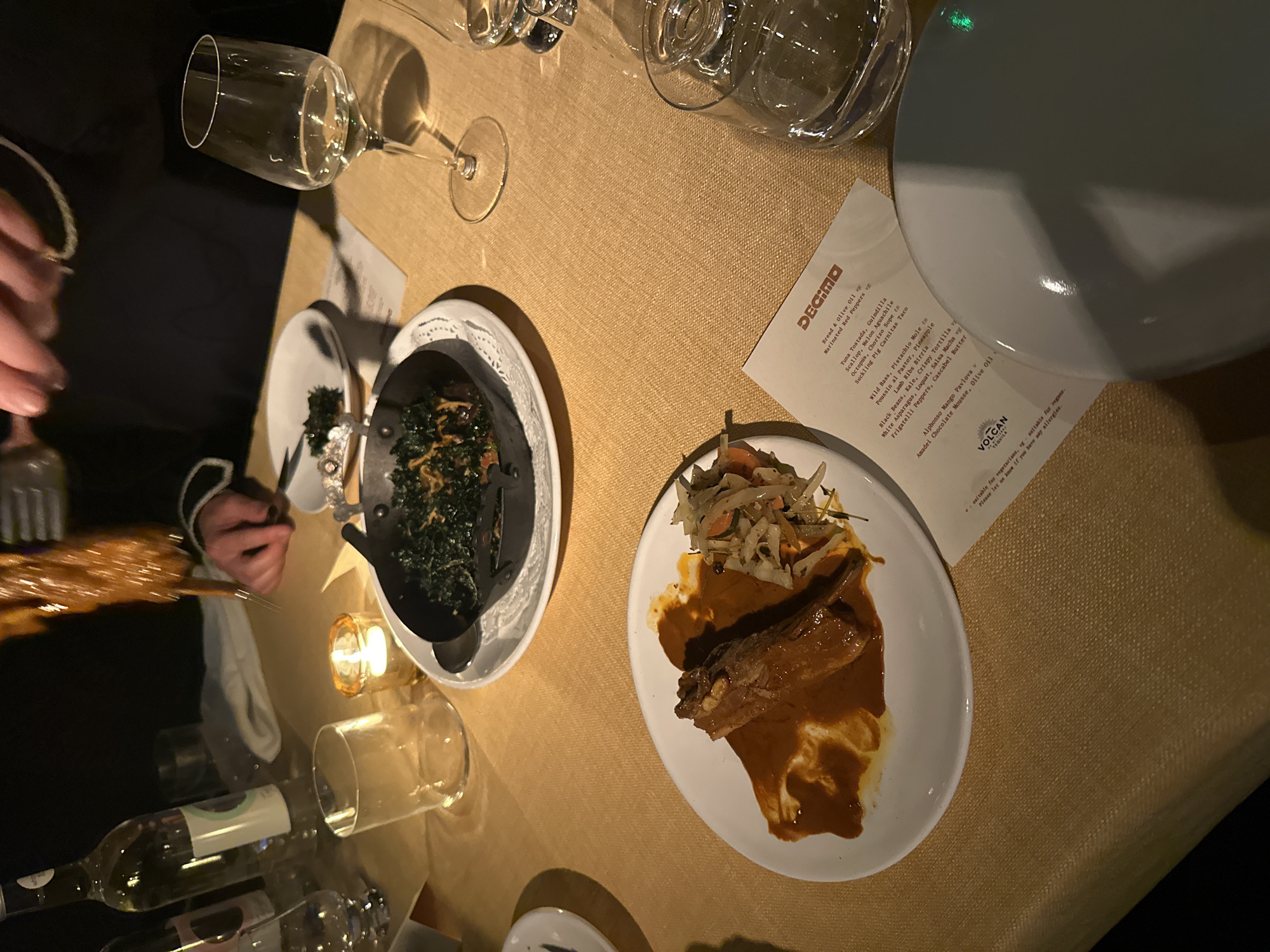The image captures a table setting at Decimo, a restaurant in London, United Kingdom. The table, covered with a textured beige cloth, holds several dining elements.
A white plate features a piece of meat covered in a rich, reddish-brown sauce, accompanied by a small slaw. Another white plate contains what appears to be sautéed greens served in a small black skillet.
Visible beverages include wine glasses, some containing liquid. A bottle of wine is seen in the bottom left corner. A folded menu for Decimo, with various dishes listed, lies on the table. A lit candle in a small glass holder provides subtle lighting.
A person, partially visible on the left, is holding a skewer with food. The scene suggests a dining experience at the restaurant, with the focus on the presented dishes and drinks.