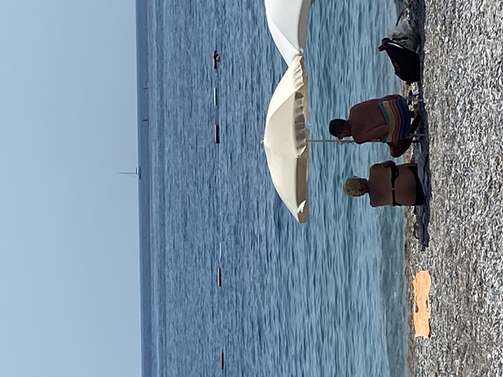 An elevated, medium shot captures a serene beach scene in Prijevor, Montenegro, under a clear, pale blue sky. The vast expanse of the Adriatic Sea dominates the upper two-thirds of the frame, its surface rippled with gentle waves and marked by a distant sailboat and two indistinct figures on what appear to be paddleboards. A line of red buoys is visible in the middle distance.
In the foreground, a couple sits on a pebble beach. A woman with blonde hair, wearing a black bikini, is seated facing the sea. Beside her, a man with dark hair, wearing sunglasses and a towel draped over his lap in a rainbow pattern, sits in a portable chair, also facing the water. They are partially shaded by a large, cream-colored beach umbrella. To the right of the man, a black backpack and a lighter colored bag are placed on the ground. An orange towel lies on the beach to the left of the woman. The water's edge is a thin line of gentle surf breaking on the pebbles.