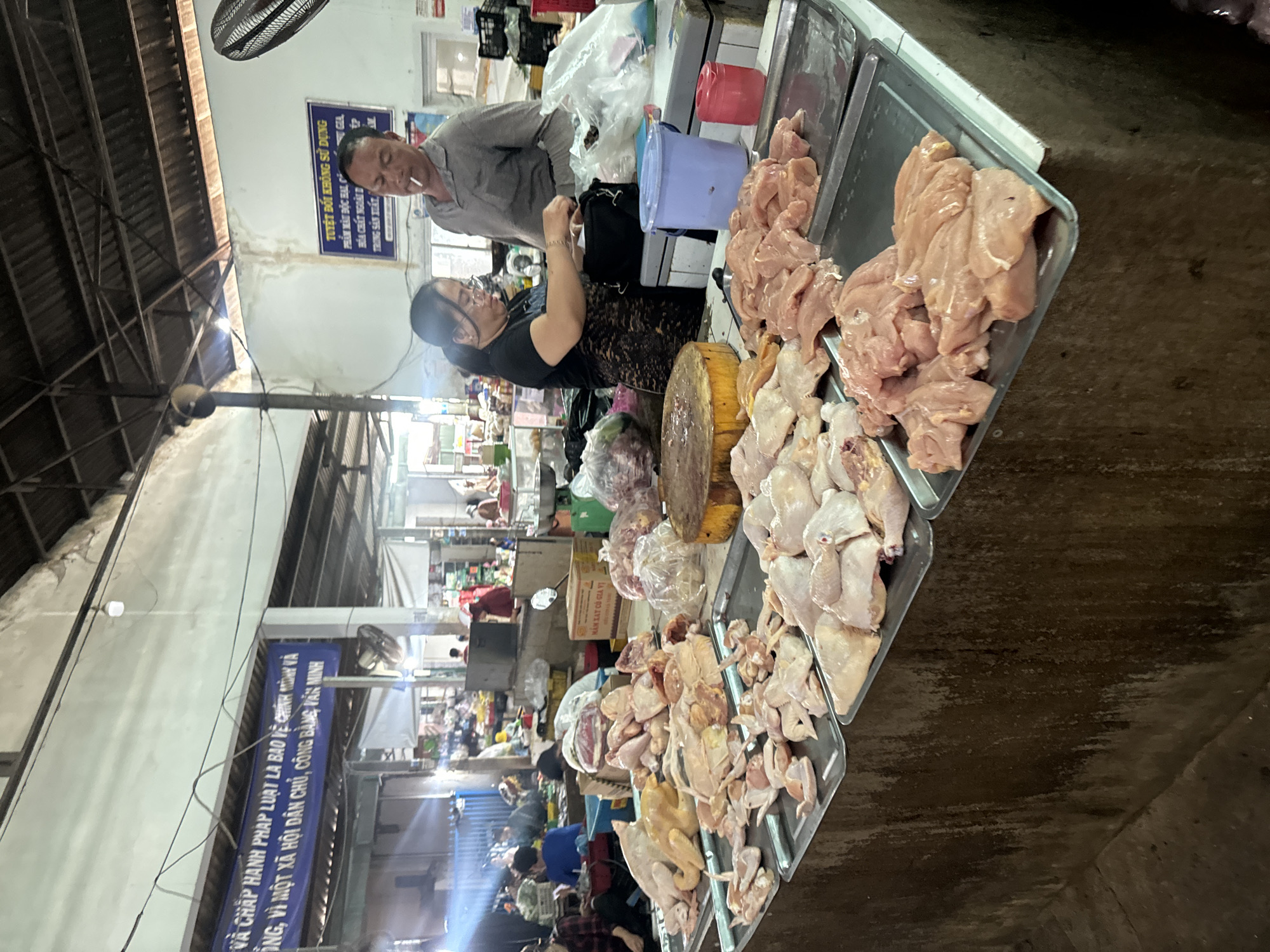 This is a medium shot photograph of a bustling indoor market scene in Mũi Né, Vietnam. In the foreground, a long, dark, wooden counter displays trays of raw chicken meat. The chicken is arranged in various cuts, some whole birds, others pre-portioned pieces, and some are still whole chickens, with their skin still attached. Behind the counter, two individuals are visible. A woman wearing glasses and a black top is sorting through a black bag. To her right, a man is standing, smoking a cigarette and wearing a grey shirt.
The background reveals the typical elements of an open-air market with a corrugated metal roof structure. Various stalls are set up, with goods and people milling about. Signs are visible on the walls, some with text. The overall atmosphere suggests a busy, commercial environment. The lighting is a mix of natural and artificial, with some areas brighter than others, indicating it might be a covered market space.