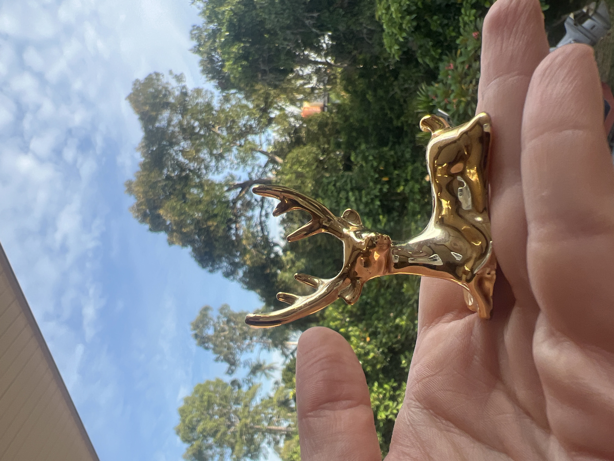 A close-up, eye-level shot shows a person's hand holding a small, shiny golden deer figurine. The deer is depicted in a seated position with its head turned to the left, its antlers prominently displayed. The hand, with visible lines and creases, cradles the figurine from below. In the background, a lush green landscape of trees and foliage stretches out under a bright blue sky with scattered white clouds. A small, orange-roofed structure is partially visible amongst the trees. The setting appears to be outdoors, possibly on a balcony or porch, suggesting a location in Ko Samui District, Thailand, given the tropical vegetation.