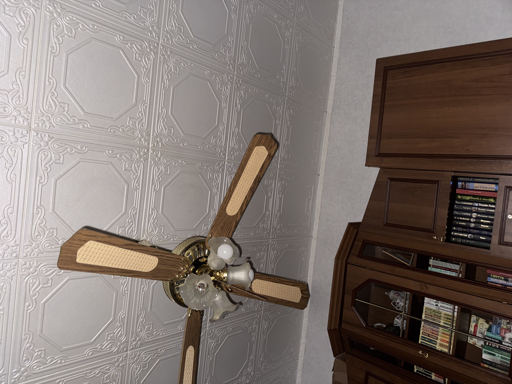 An indoor, eye-level, medium shot captures a close-up view of a ceiling fan with three wooden blades, each adorned with a beige insert, installed on a white embossed ceiling. The fan also features a brass-colored base and three glass light fixtures, two of which contain visible light bulbs. Below the ceiling, the top portion of dark brown wooden cabinetry with glass-fronted shelves displaying books and decorative items is visible. The scene is lit by the overhead lights and ambient room lighting, creating soft shadows. The image is consistent with an interior space in a residential or commercial building.