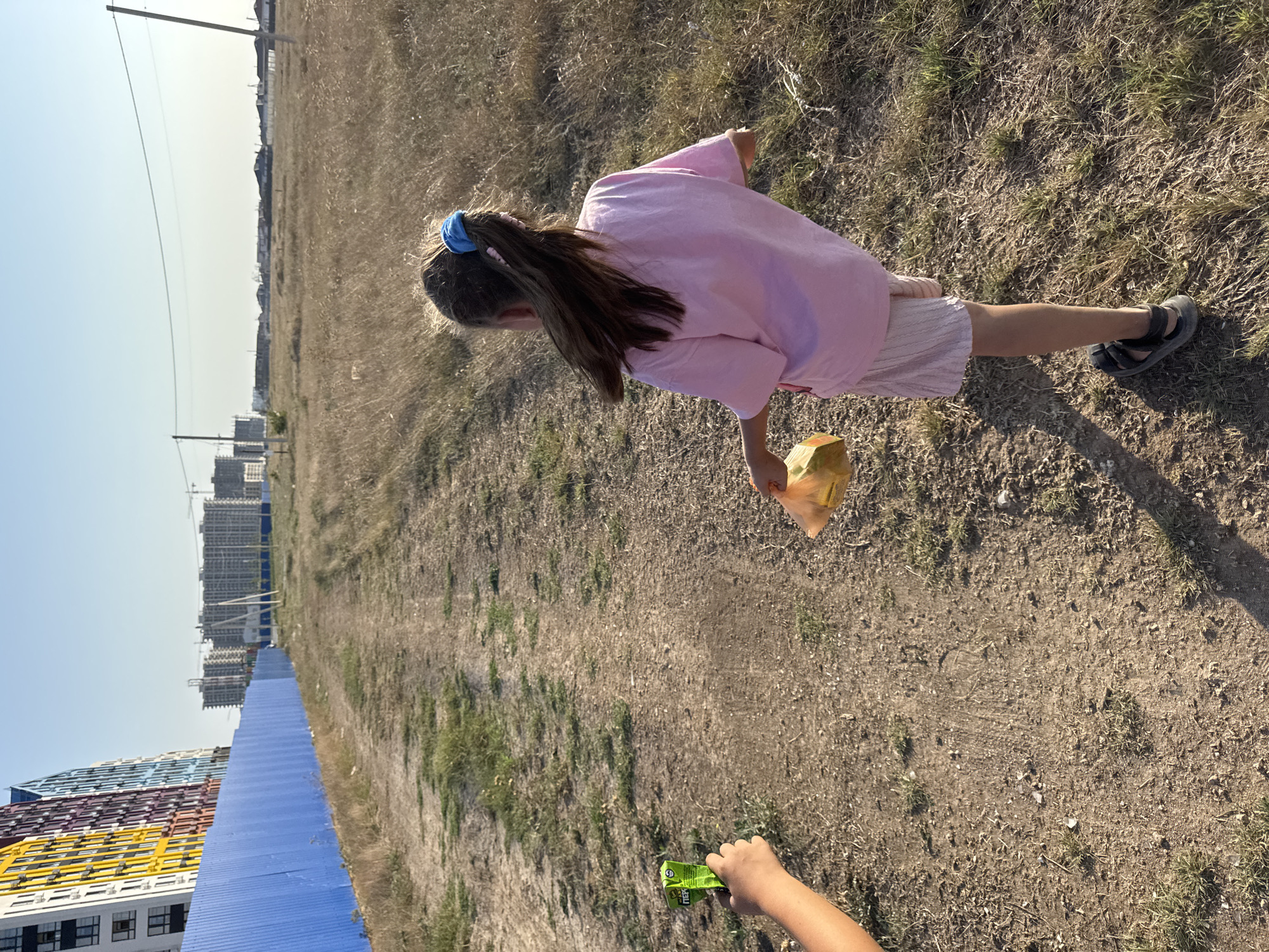 A girl with brown hair in a ponytail, wearing a pink t-shirt and a light-colored skirt, is walking away from the camera on a dirt path. She is holding a crumpled yellow bag in her right hand. To the left of the path, a person's arm is visible, holding a green package with Cyrillic text. The background shows a bright blue fence, a row of colorful apartment buildings, and construction sites with cranes. The ground is covered in dry grass and dirt, suggesting an arid environment. The sky is clear and blue. The scene appears to be in Yevpatoriya, Ukraine, a city known for its coastal location and developing infrastructure.