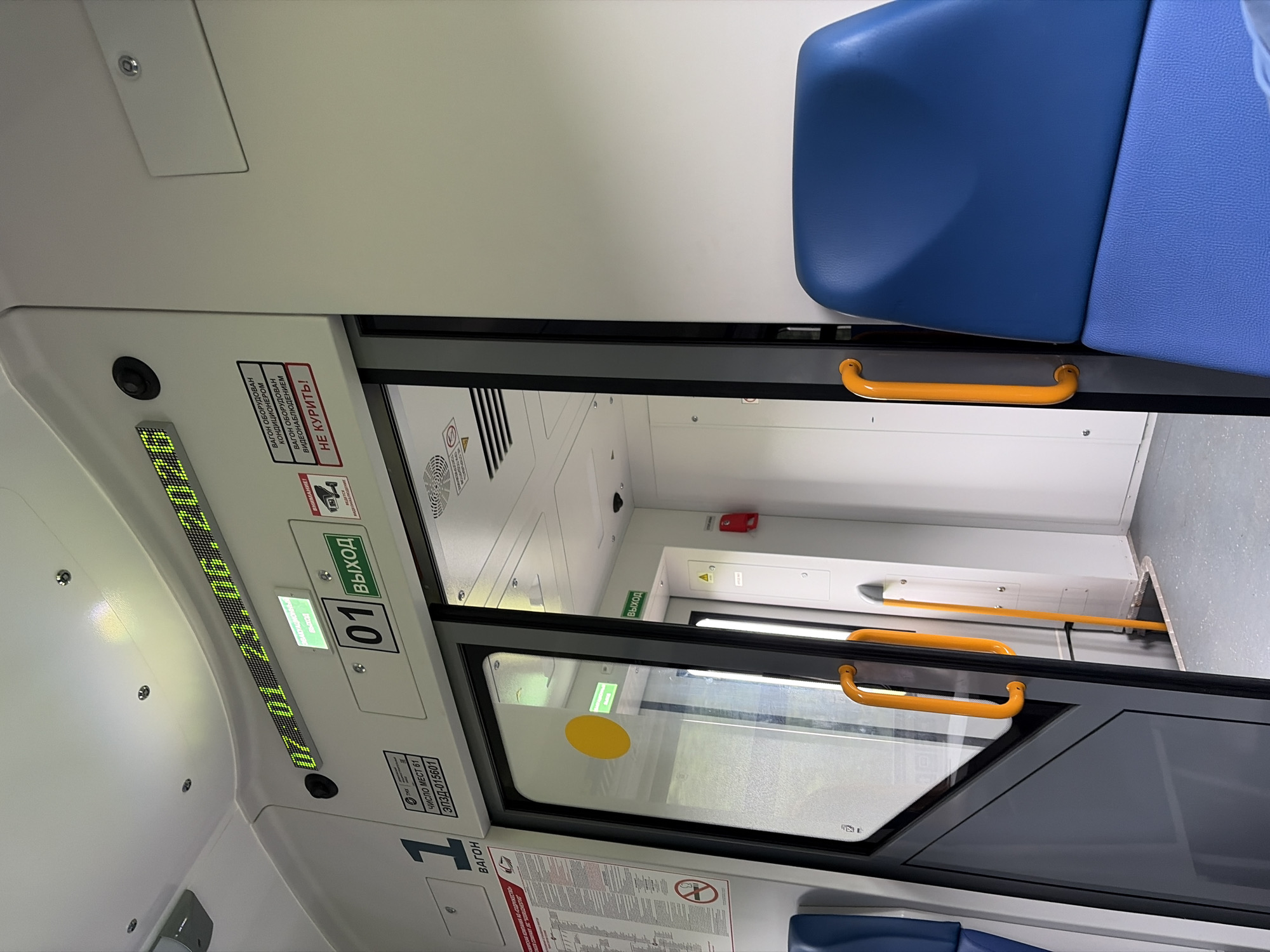 The image captures the interior of a passenger train car. In the foreground, a blue seat is visible on the right. A closed sliding door with orange handles is on the left, revealing a glimpse of the interior beyond. Above the door, a digital display shows the date and time: "07:01 23.06.2026". Adjacent to the time display are signs indicating "АВАРИЙНЫЙ ВЫХОД" (Emergency Exit) and "01 ВЫХОД" (01 Exit). Further to the right, there are smaller signs, one with a camera icon and text that appears to be a warning about video surveillance, and another red sign with "НЕ КУРИТЬ!" (Do not smoke!). To the left of the door, a sign with the number "1" and the word "ВАГОН" (Car) is prominently displayed. The overall environment suggests a clean and modern train interior. The prompt mentions the city of Баграш-Бигра, Russia, but this information is not visually verifiable from the image itself.