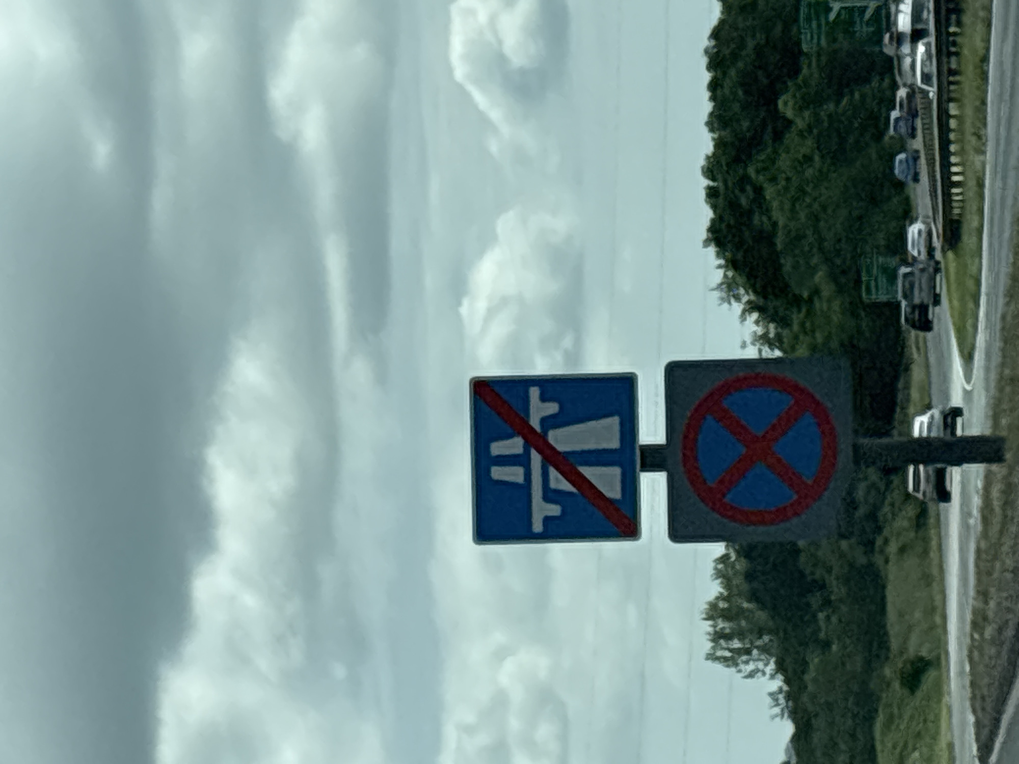 The image shows two road signs, one prohibiting vehicles carrying water-polluting substances and the other indicating "no stopping." A cloudy sky fills the background, while roadside trees and parked vehicles are visible on the right. The location appears to be near a road outside of the city of Chester, United Kingdom.