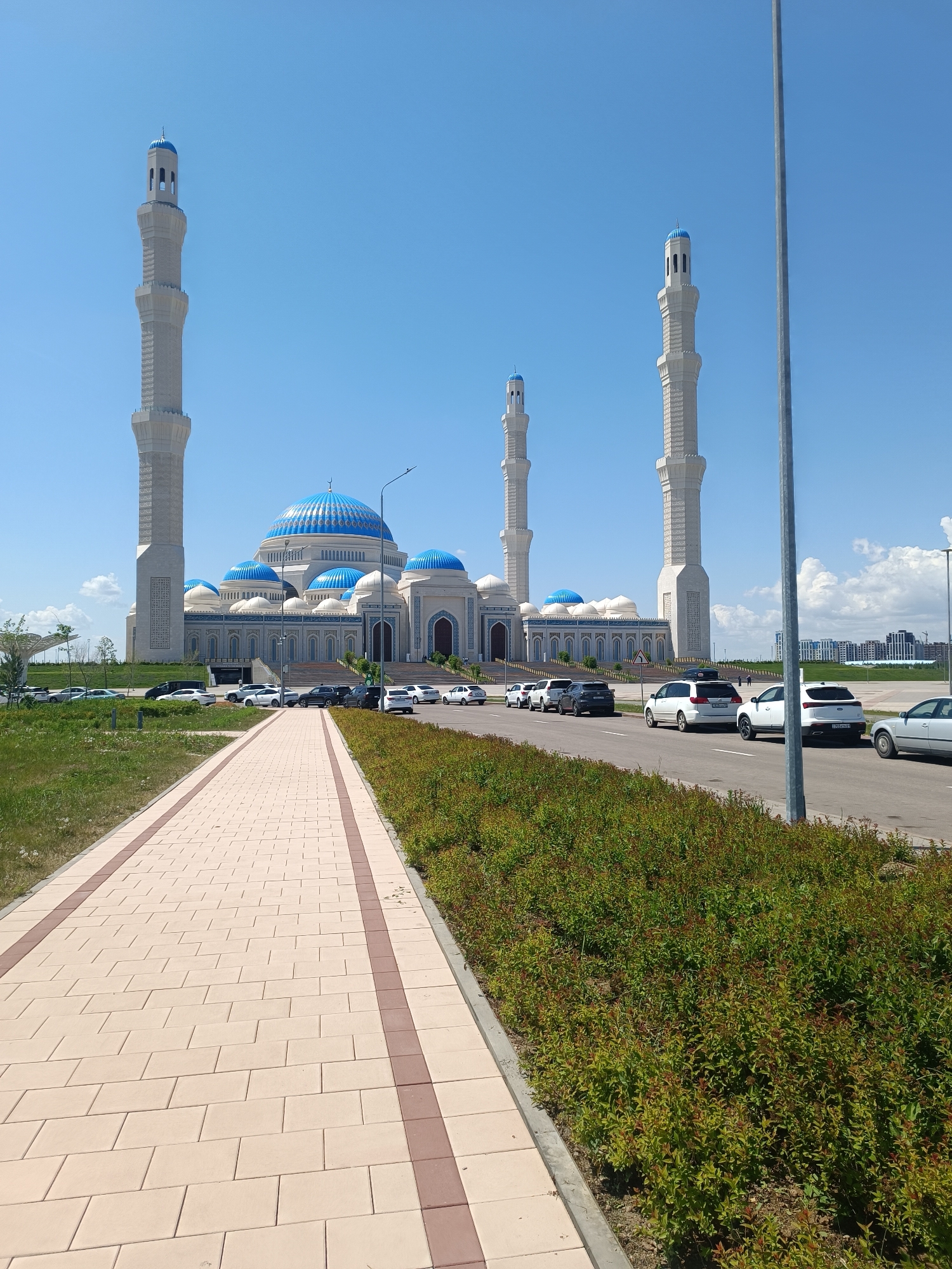The image shows the Hazrat Sultan Mosque in Astana, Kazakhstan. A paved walkway leads towards the mosque. A line of cars is parked along the road in front of the mosque. The sky is blue with some clouds. The mosque has white minarets with blue domes. There are some trees and bushes to the left and right of the walkway.