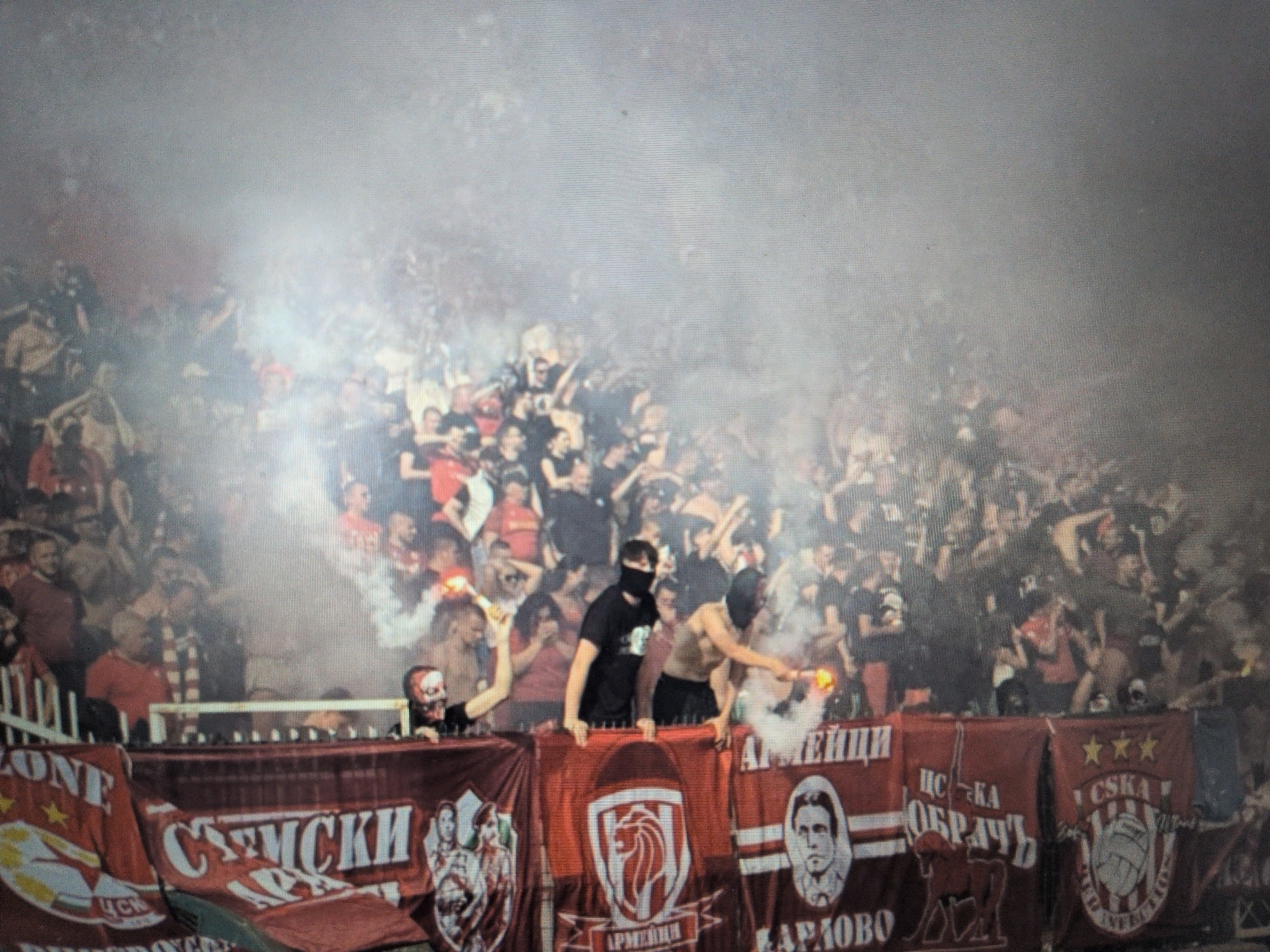 The image captures a highly energetic and smoky scene at what appears to be a night-time sporting event, likely a football match in a stadium. A large crowd of people, predominantly young men, are densely packed in the stands, many with their arms raised, seemingly cheering or chanting.
The primary activity is a passionate display of fan support, characterized by the lighting of multiple flares and pyrotechnics. These devices emit intense bright light and thick plumes of white and reddish smoke, which rise into the air, partially obscuring the upper sections of the crowd and creating a hazy, dramatic atmosphere.
In the foreground, several individuals stand on or behind a metal barrier, directly engaging in the pyrotechnic display. Three men are particularly prominent: one on the left wears a skull-like face mask and holds a lit flare; a central figure in a black t-shirt has a balaclava covering his face; and another individual to the right, bare-chested, is also lighting a flare, contributing to the dense smoke cloud.
Notable details include the dramatic illumination provided by the flares, casting an orange and red glow on the faces of some spectators and the smoke itself. The scene is clearly nocturnal, given the effectiveness of the artificial light sources.
Along the bottom of the image, several large banners are displayed on the barrier, predominantly in red and white. Visible text in Cyrillic script provides significant context:
* "ZONE" and "ЦСКА" (CSKA) appear on the left.
* "СЕМСКИ" is visible on a banner to the left of the center.
* A central banner features a crest with a lion or similar animal head and the word "АРМЕЙЦИ" (Armeitsi), referring to "Army Men."
* Another banner displays a portrait of a man (likely Vasil Levski, a Bulgarian national hero) above "ЦСКА ОБРАЧЪ" and "КАРЛОВО" (Karlovo, his birthplace).
* The far-right banner shows three stars, "ЦСКА", and a crest incorporating a basketball within a star/circle, strongly identifying the supporters as fans of CSKA Sofia, a Bulgarian sports club.