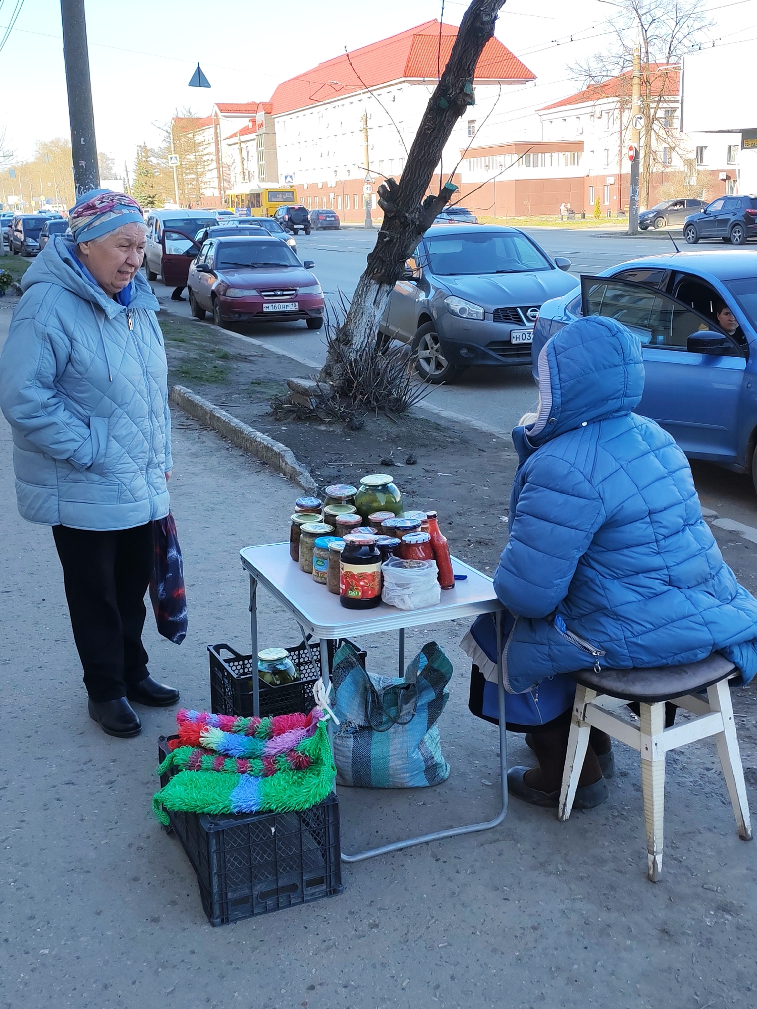 The image captures an outdoor street scene in Izhevsk, Russia, featuring two elderly women and a makeshift street stall. On the left, a woman stands, facing right, wearing a light blue quilted jacket, dark trousers, and black shoes. Her hands are in her pockets, and she has a colorful patterned headscarf. She appears to be speaking, with her mouth slightly open. A patterned bag hangs from her left hand.
To the right, another woman is seated on a simple wooden stool, facing away from the camera. She wears a dark blue quilted hooded jacket and brown boots. She is positioned behind a small, white, foldable table laden with various glass jars, likely containing homemade preserves or pickled goods. Notable items on the table include several jars of green pickles, a jar with red contents (possibly tomatoes or sauce), and a bottle of red sauce.
Beneath the table, two black plastic crates are visible. One crate holds a bundle of brightly colored, fluffy scrubbers or sponges in shades of green, blue, pink, and red. The other crate contains a single large jar of pickles. A striped woven bag rests on the ground next to the crates.
The setting is a paved sidewalk alongside an asphalt road. In the background, numerous cars are parked along the curb and driving on the street. Visible license plates include "M 160 HP 18" on a burgundy sedan and a partially visible "H 03" on a grey Nissan Qashqai, with "18" indicating the Udmurt Republic where Izhevsk is located. Beyond the road, large buildings with red roofs and white/brown facades line the street. A pedestrian crossing sign and a small "40" speed limit sign are discernible in the distance. The weather appears cool, as indicated by the heavy jackets worn by both women, under bright daylight. The scene suggests a casual exchange, possibly a purchase, at a roadside vendor setup.