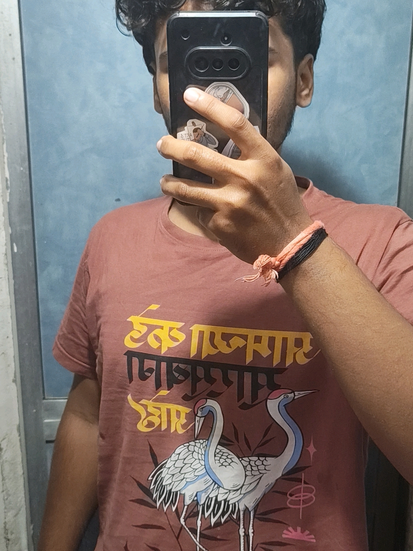 The image is a close-up, waist-up selfie of a young South Asian male, taken indoors. He is wearing a maroon t-shirt with an image of two cranes and yellow and black text in an Indian script. A black and orange woven bracelet is visible on his right wrist. His face is partially obscured by a black smartphone he is holding to take the picture. The background appears to be a blue wall, possibly a door or partition, with a vertical white element on the left side. The lighting suggests an indoor setting, likely a home or private space. No specific details link this image to the city of Pune, India.