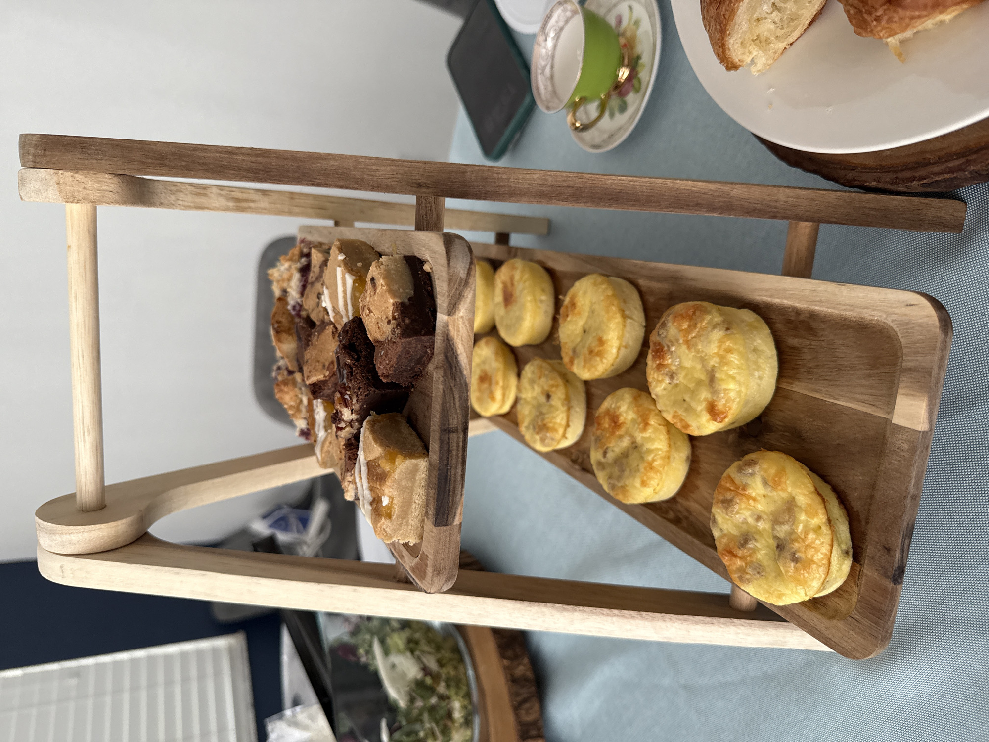 The image depicts a tiered wooden serving tray filled with assorted pastries and baked goods, possibly arranged for a small gathering or afternoon tea. The tray features a rectangular shape with multiple levels, supported by vertical posts. The bottom level holds a selection of bite-sized cakes and brownies, while the top level displays a row of savory scones.
In the background, a light blue tablecloth covers the table surface. Other items visible on the table include a teacup and saucer with a floral pattern, a partially visible smartphone, a plate with croissants, and a salad bowl.
The location is the city of Davie, United States of America.