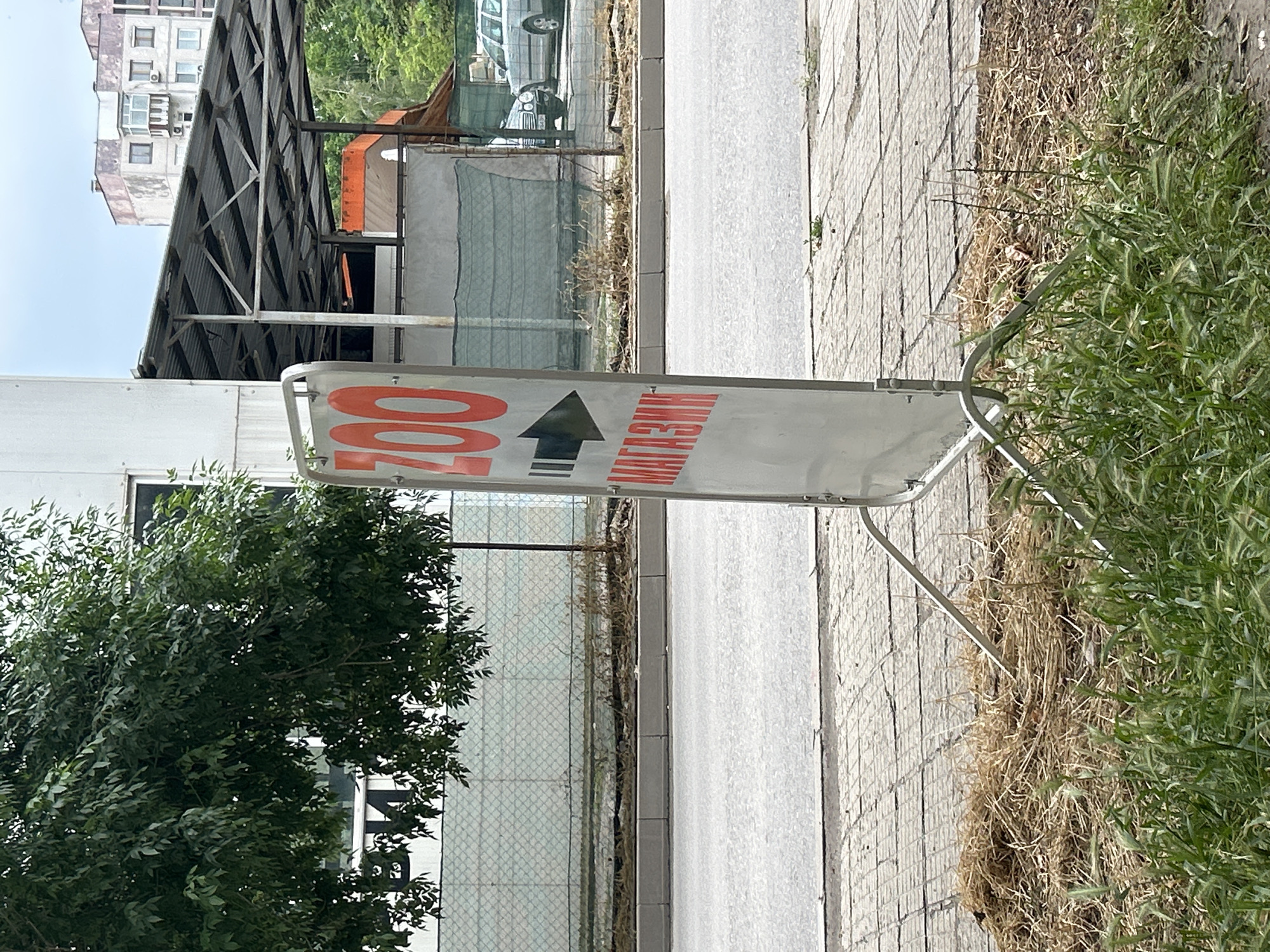 This outdoor daytime image captures an A-frame sign on the side of a paved path, directing viewers towards a destination. The sign is white with prominent orange Cyrillic text and a black arrow. On the left side of the sign, the letters "ZOO" are visible in large orange characters. A thick black arrow points upwards in the center, and to the right, the word "МАГАЗИН" (MAGAZIN), which translates to "shop" or "store," is written in orange Cyrillic letters. This indicates a "ZOO shop" is located in the direction of the arrow.
The paved path, possibly a sidewalk or a small road, is bordered on one side by patches of green grass and dry, straw-like weeds. A lush green tree with dense foliage is situated to the left of the paved area, with some of its leaves obscuring a small part of a background structure.
In the background, a variety of buildings and structures are visible under a clear, bright sky. These include a white, possibly modular or trailer-like building, and behind it, a large, dark corrugated metal roof structure, perhaps part of a commercial or industrial facility. Further back, an older, multi-story residential building with several windows and balconies can be seen. A fence is also present in the middle ground, separating different areas. The presence of Cyrillic text aligns with the location context of Plovdiv, Bulgaria. The bright lighting suggests it is daytime, and the dry grass might indicate a warm season.