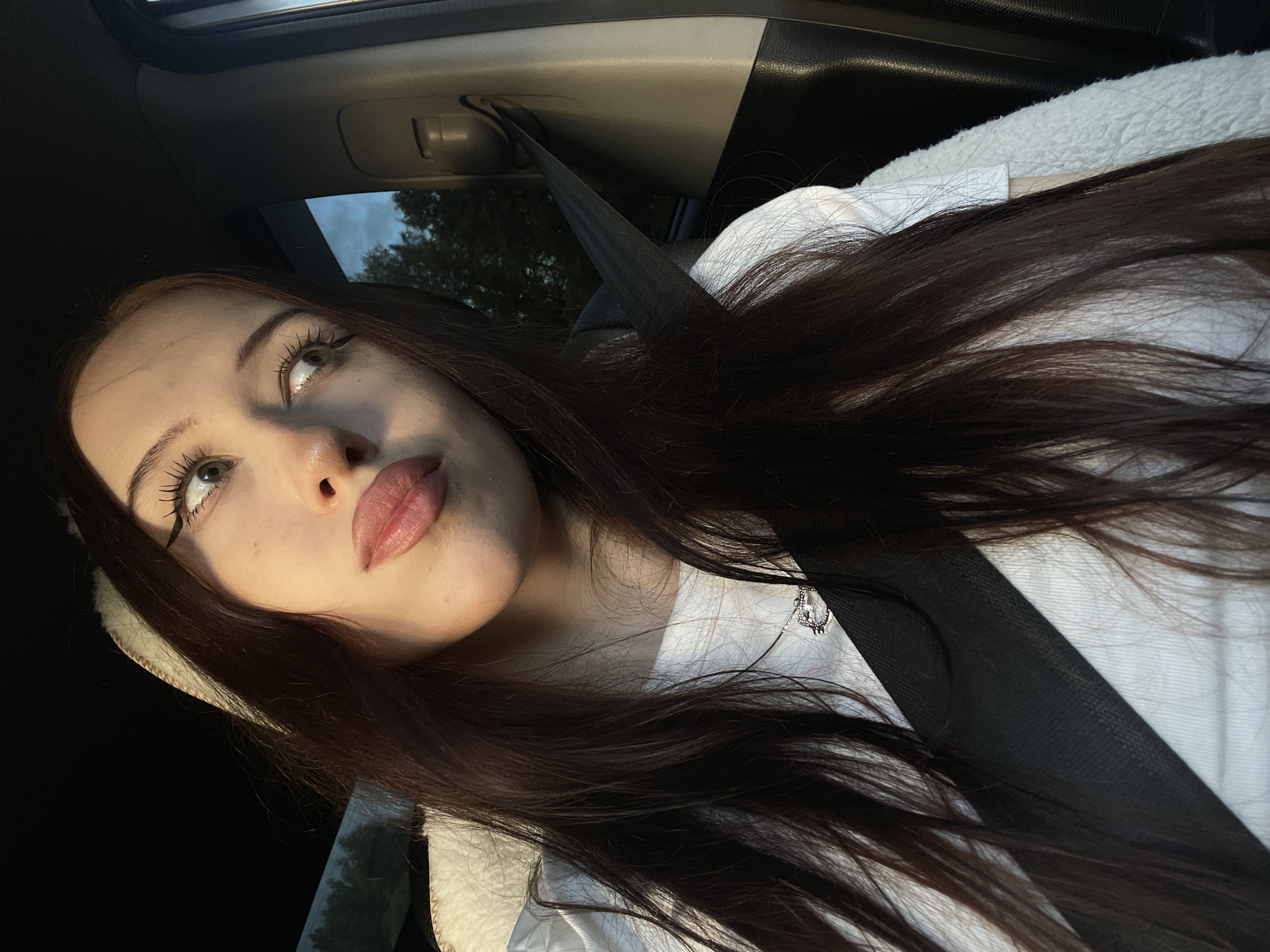 In this close-up, low-angle selfie, a young woman with long, dark brown hair is captured from the chest up, inside what appears to be a car. The woman is wearing a white top and a fluffy white hooded jacket. A black seatbelt crosses diagonally over her body, and a silver necklace with a small pendant is visible. Her makeup includes winged eyeliner and lip gloss. She is looking upwards and to her left, with a slight smile.
The setting suggests a daytime scene, possibly late afternoon or early evening, due to the warm lighting casting shadows and highlighting her features. Through the car window, trees and a cloudy sky are visible, indicating an outdoor location, likely in a rural or suburban area given the presence of trees and the caption's location context of Gavrilovka, Russia. The overall mood is relaxed and contemplative, perhaps reflecting a moment of quiet reflection during a drive. No visible text is present in the image.