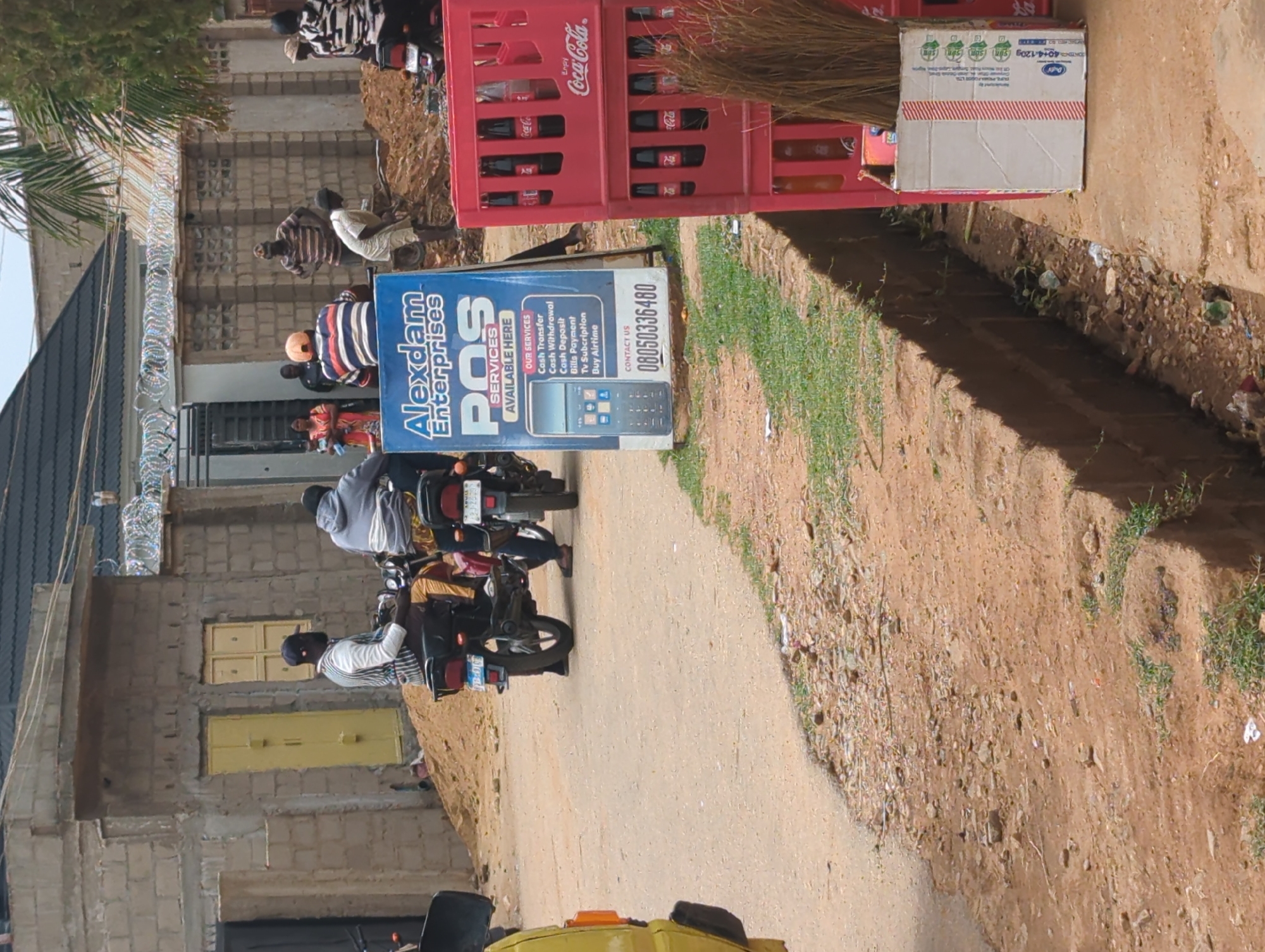 The image shows a street scene in Sabon Tasha, Nigeria. A Coca-Cola crate and a promotional sign for "Alexdam Enterprises POS Services" are visible. People are riding motorcycles on the street. Buildings are visible in the background. The ground is dirt and there are small patches of vegetation.