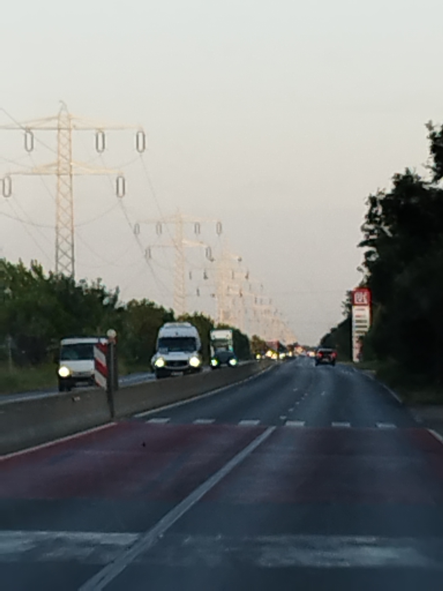 The image shows a highway in Gulia, Romania, with multiple vehicles and high-voltage power lines. The scene appears to be captured from a vehicle moving on the highway. In the foreground, the road surface is visible, marked with red and white stripes, possibly indicating a pedestrian crossing or a safety zone. A concrete barrier separates the lanes of traffic.
Several vehicles are present on the highway. In the middle ground, there are at least three vans or small buses, two of which are white and one appears to be light-colored. Their headlights are on, suggesting it might be dusk or overcast. Further down the road, more vehicles are visible, including a dark-colored car and other indistinct vehicles.
On the left side of the highway, there are trees and vegetation. Towering over the landscape are numerous high-voltage power line towers, a common sight in rural and industrial areas. On the right side of the highway, a tall, red and white signpost, possibly a gas station advertisement, is visible among the trees. The sky is overcast, creating a diffused light. The overall impression is one of a typical highway scene, with elements of infrastructure and transportation.