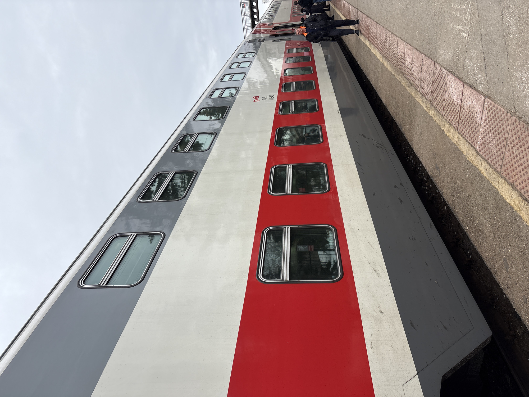A bi-level passenger train is stopped at a concrete platform under an overcast sky in Dimitrovgrad, Russia. The train car is striped horizontally in grey (top), white (upper middle), red (lower middle), and dark grey (bottom). The "РЖД" logo and the text "065 144-40" are visible on the white section. Multiple rectangular windows with rounded corners are present on both levels, reflecting the environment. The platform features a reddish-brown tactile paving strip and a yellow safety line. In the immediate foreground, a man, wearing a dark jacket with an orange lining and a dark cap, walks on the platform parallel to the train, facing left. Further down the platform to the right, other individuals are visible near an open train door, consistent with passenger activity. A metal pedestrian overpass is partially visible in the distant background above the train.