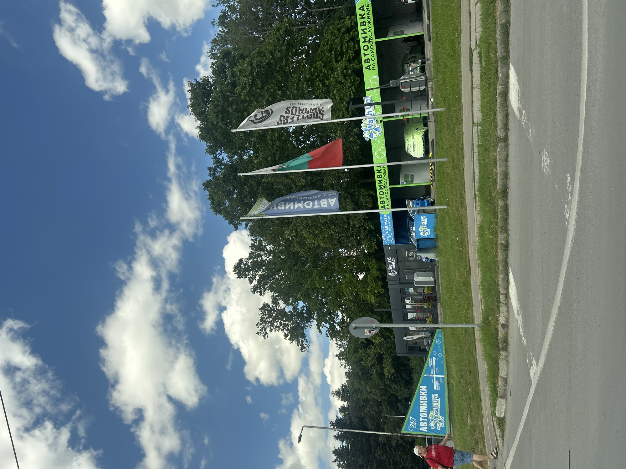 The image captures a self-service car wash facility in Varna, Bulgaria, on a bright, sunny day with scattered white clouds against a blue sky. The car wash is branded with signs in Bulgarian, including "Автомивки" (Car Wash) and "НА САМООБСЛУЖВАНЕ" (Self-Service). Three flags are prominently displayed, one being the Bulgarian national flag, and the other two with logos and text related to car detailing and washing services. Several cars are present at the car wash bays, with one lime green car appearing to be undergoing a cleaning process. A man in a red shirt and blue shorts is walking across a pedestrian crossing in the foreground, moving from left to right. A street lamp and trees line the background, indicating an outdoor urban setting. The overall scene depicts a functional commercial establishment within a city environment.