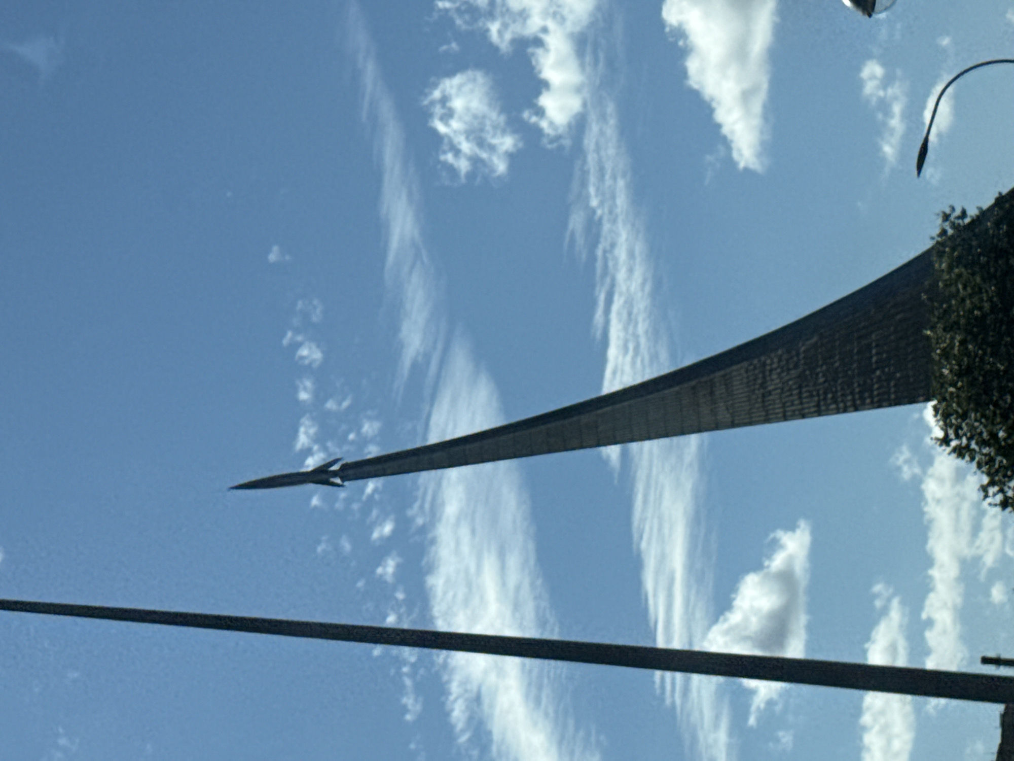 The image shows a low-angle view of a tall, pointed monument against a bright blue sky with scattered white clouds. A dark, slender pole stands to the left of the monument. The monument itself is a dark, faceted structure that tapers to a sharp point, resembling a rocket. The lower part of the monument is partially obscured by dense green foliage. A curved lamppost is visible in the lower right corner, its light fixture out of frame. The scene appears to be outdoors in a city setting. This monument is the Monument to the Conquerors of Space, located in Moscow, Russia, near the Ostankino Tower.