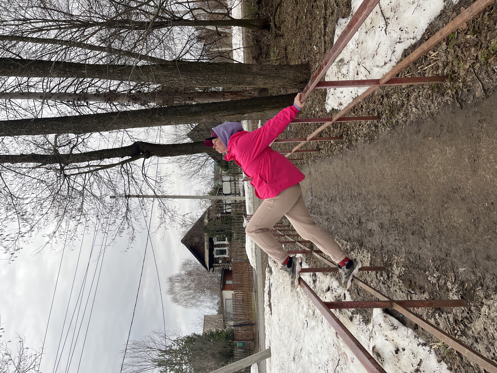 A woman in a pink jacket and tan pants is climbing over a rusty metal fence in Chaikovsky, Russia. There is snow on the ground near the fence. In the background are leafless trees and wooden houses. The sky is overcast.