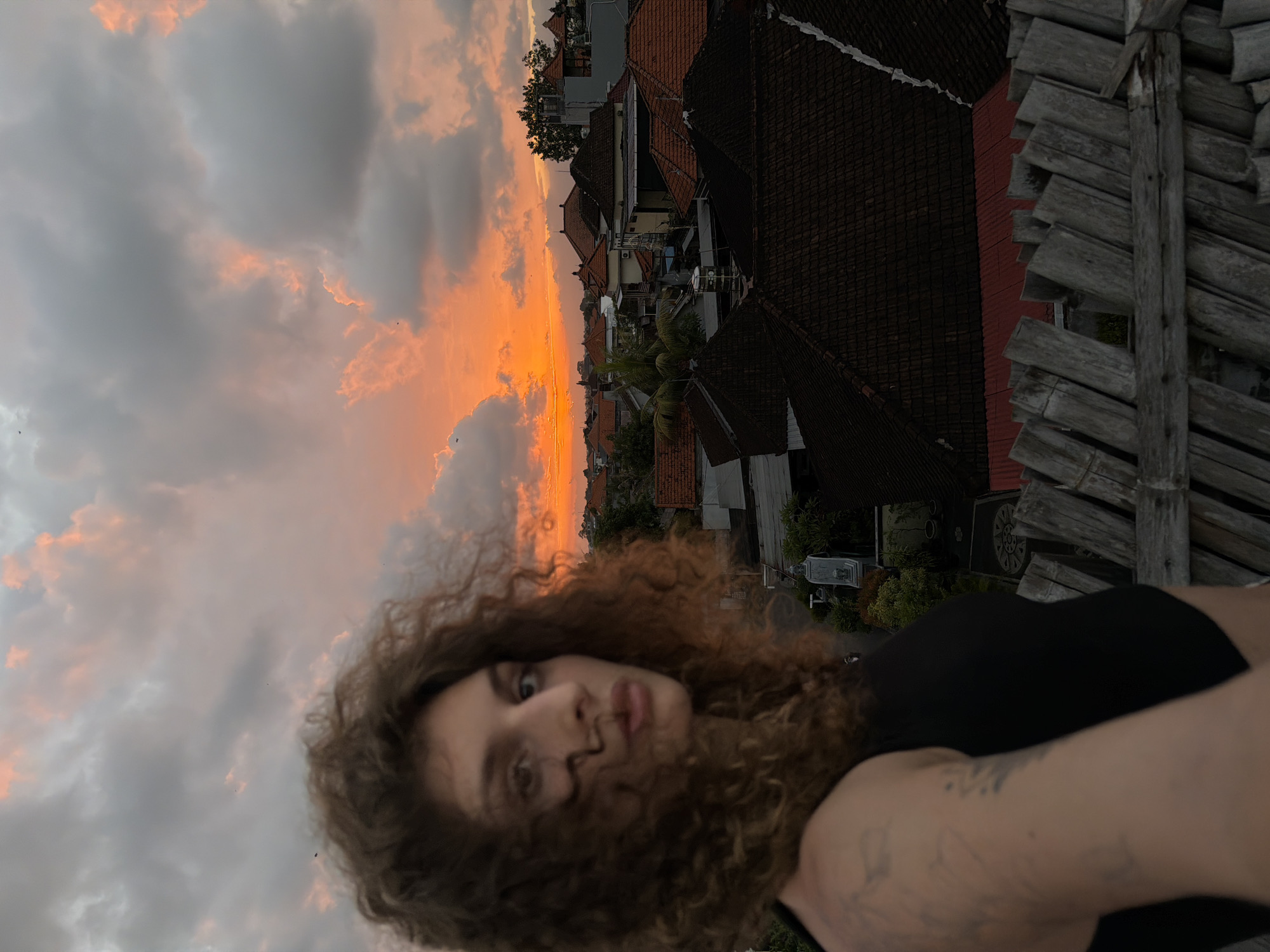 This is a portrait of a young woman with curly brown hair, captured in an outdoor setting during what appears to be sunset. The image is oriented vertically. The woman is in the foreground, with her face slightly out of focus, looking towards the camera. She is wearing a black top. Her arms are visible, with tattoos on her left arm.
The background reveals a panoramic view of a residential area, likely in North Kuta, Indonesia, given the architectural style with terracotta tiled roofs and lush greenery. The sky is dramatic, with a vibrant orange and pink sunset casting a warm glow on the clouds and the buildings. The time of day is clearly sunset, indicated by the colors in the sky. The overall mood is tranquil and atmospheric, with the natural beauty of the sunset contrasting with the urban landscape.
To the right of the frame, there is a wooden structure made of planks, possibly a fence or part of a balcony, adding a rustic element to the scene. A few small birds are visible in the sky. There is no visible text in the image.