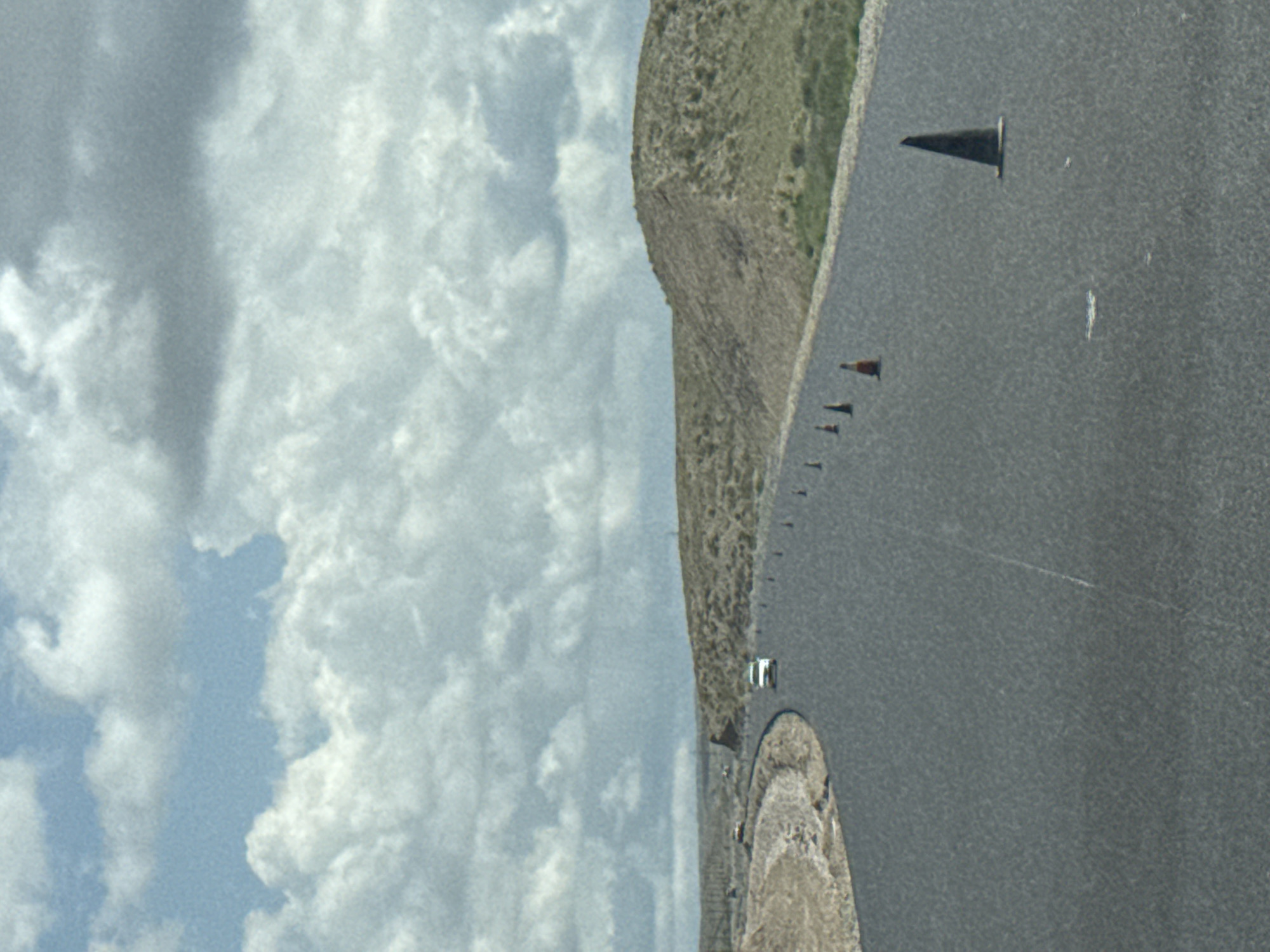 An aerial drone view captures a barren, hilly landscape under a cloudy sky. A paved road curves through the scene, with several orange traffic cones lining its edge. Two cars are visible on the road, one white and one light-colored, suggesting a travel or construction context. The terrain is sparsely vegetated, with dry grasses and shrubs covering the rolling hills. The overall impression is one of remoteness and perhaps ongoing roadwork or maintenance in a natural, undeveloped area.