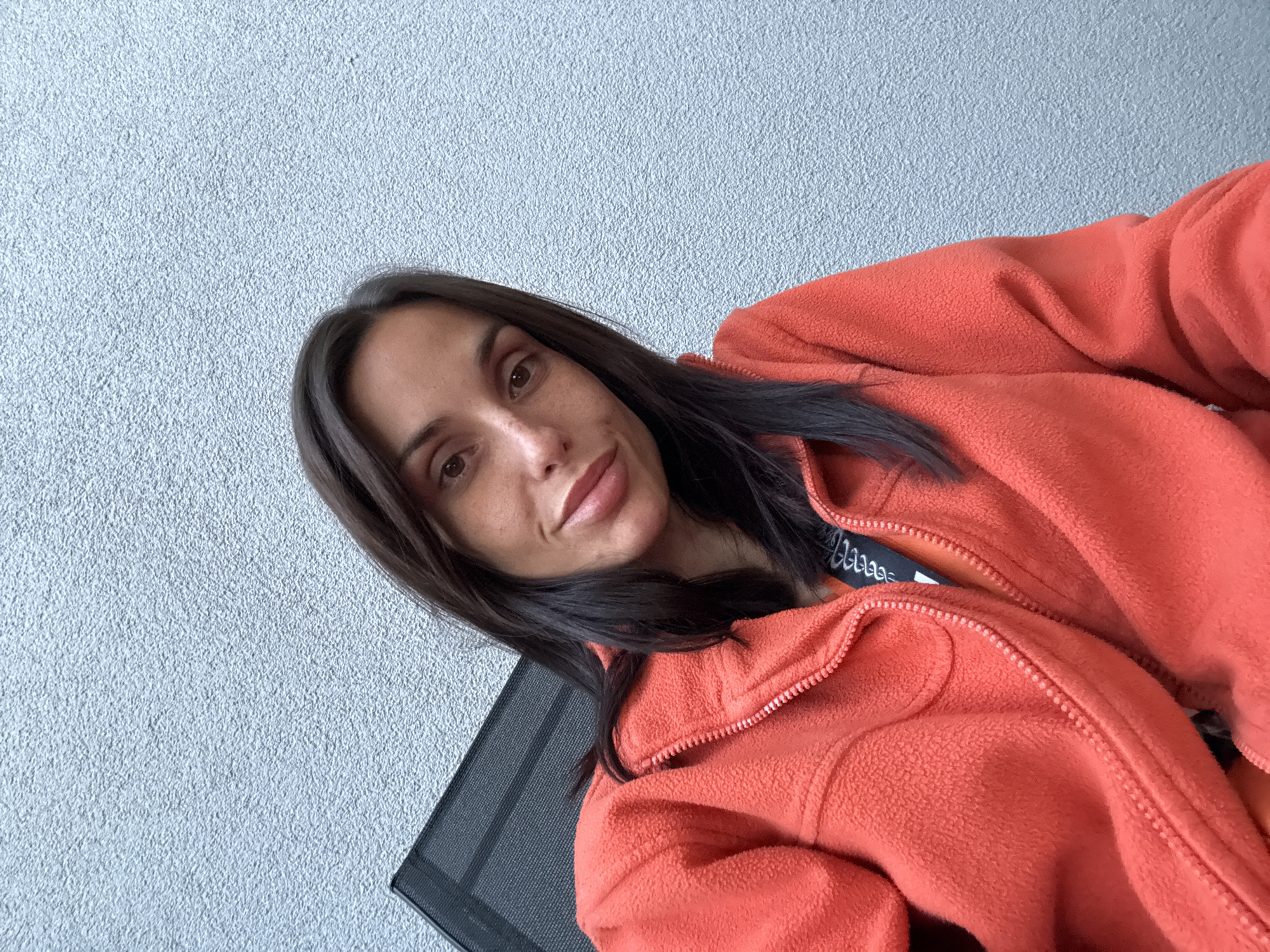 A woman with long brown hair and brown eyes is photographed at an angle, wearing an orange fleece jacket. The background is a light gray textured wall, and the edge of a dark gray mesh chair is visible. The location is Pleven, Bulgaria.