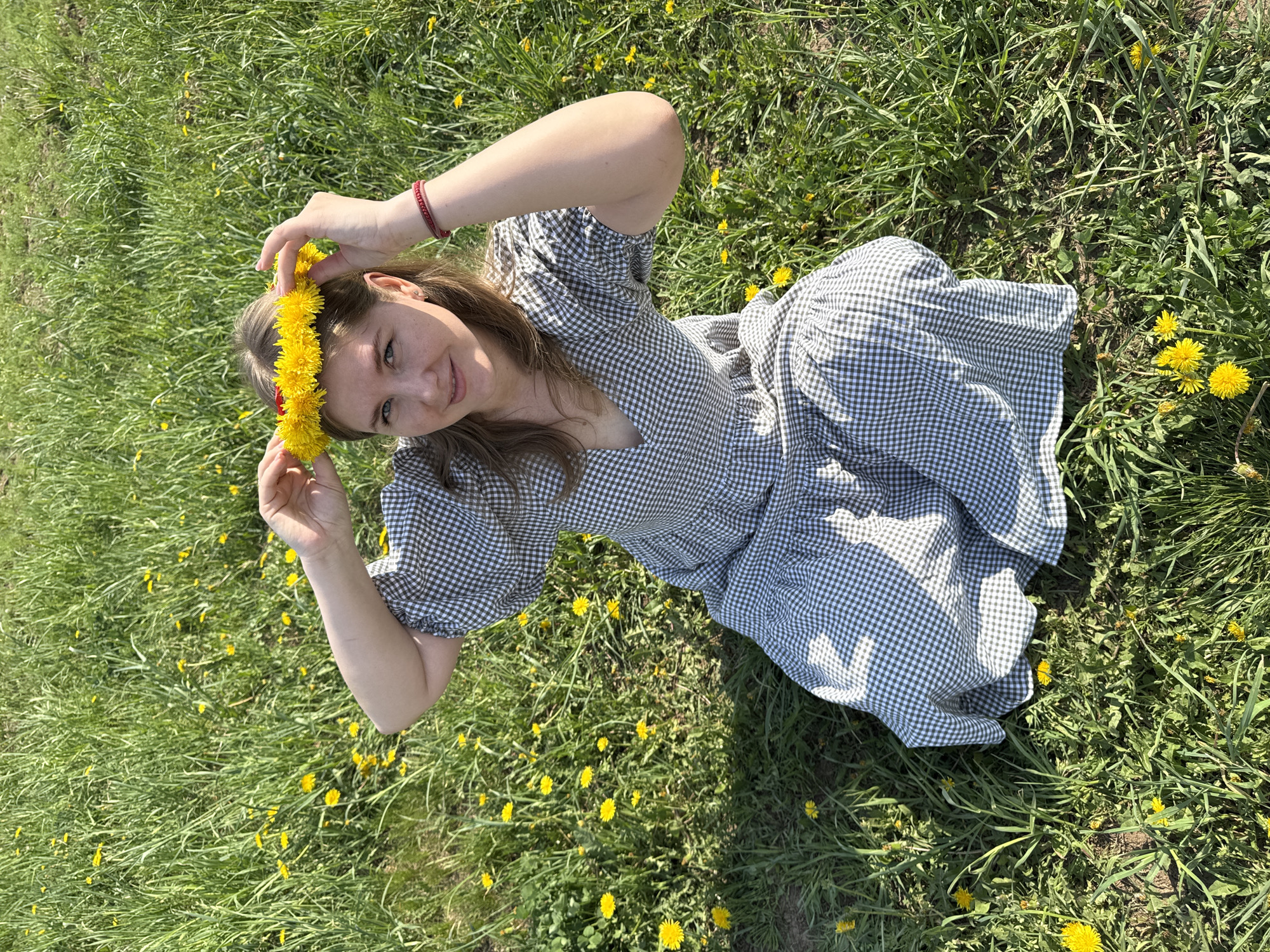 A young woman lies in a grassy field dotted with yellow dandelions, wearing a blue and white checkered dress and a yellow flower crown. She is holding the crown on her head with both hands. She is in a field in the city of Ферма, Russia.